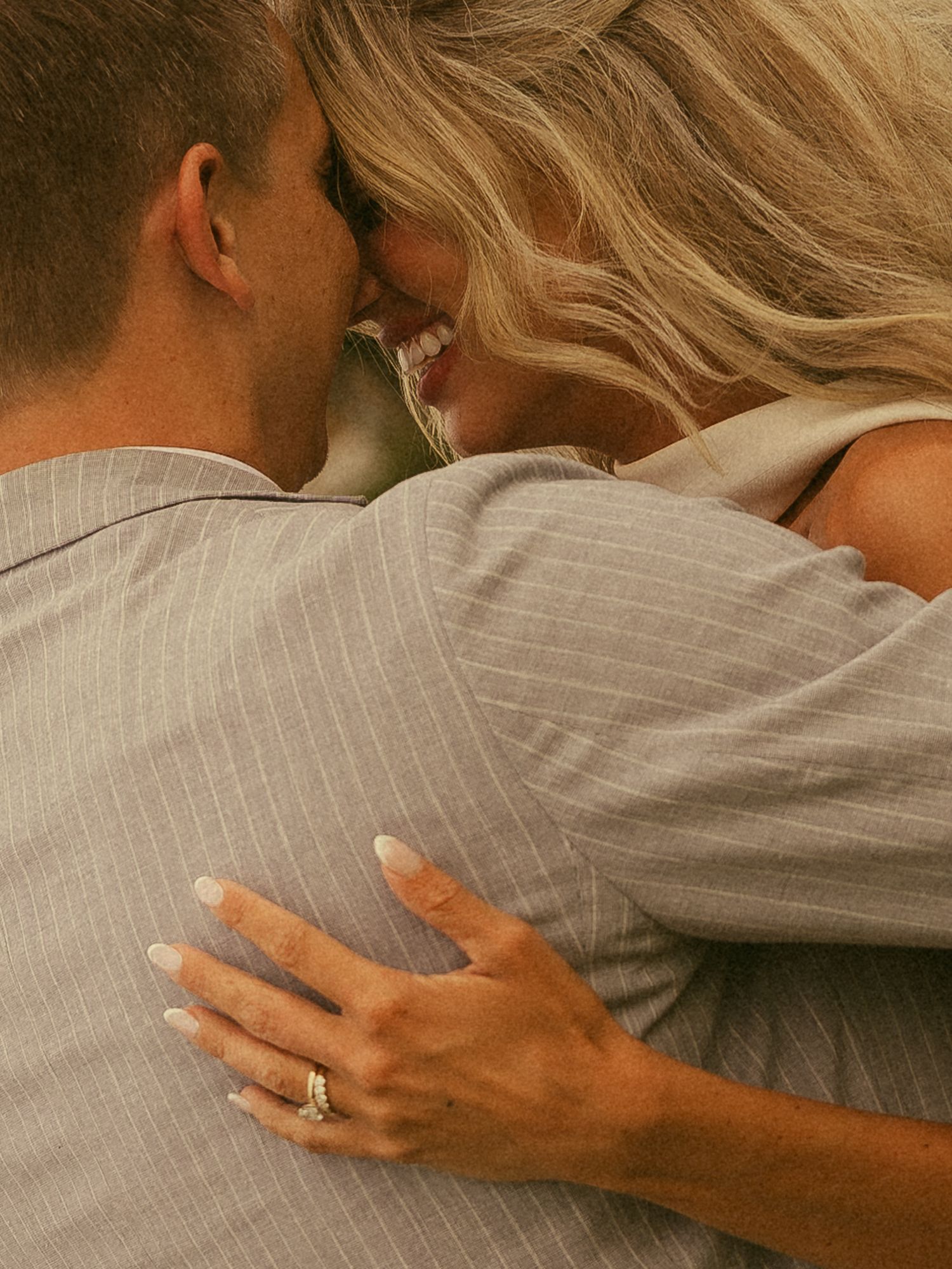 A couple in an intimate embrace, woman with blonde hair wearing a gold ring, man in a grey pinstripe suit jacket.