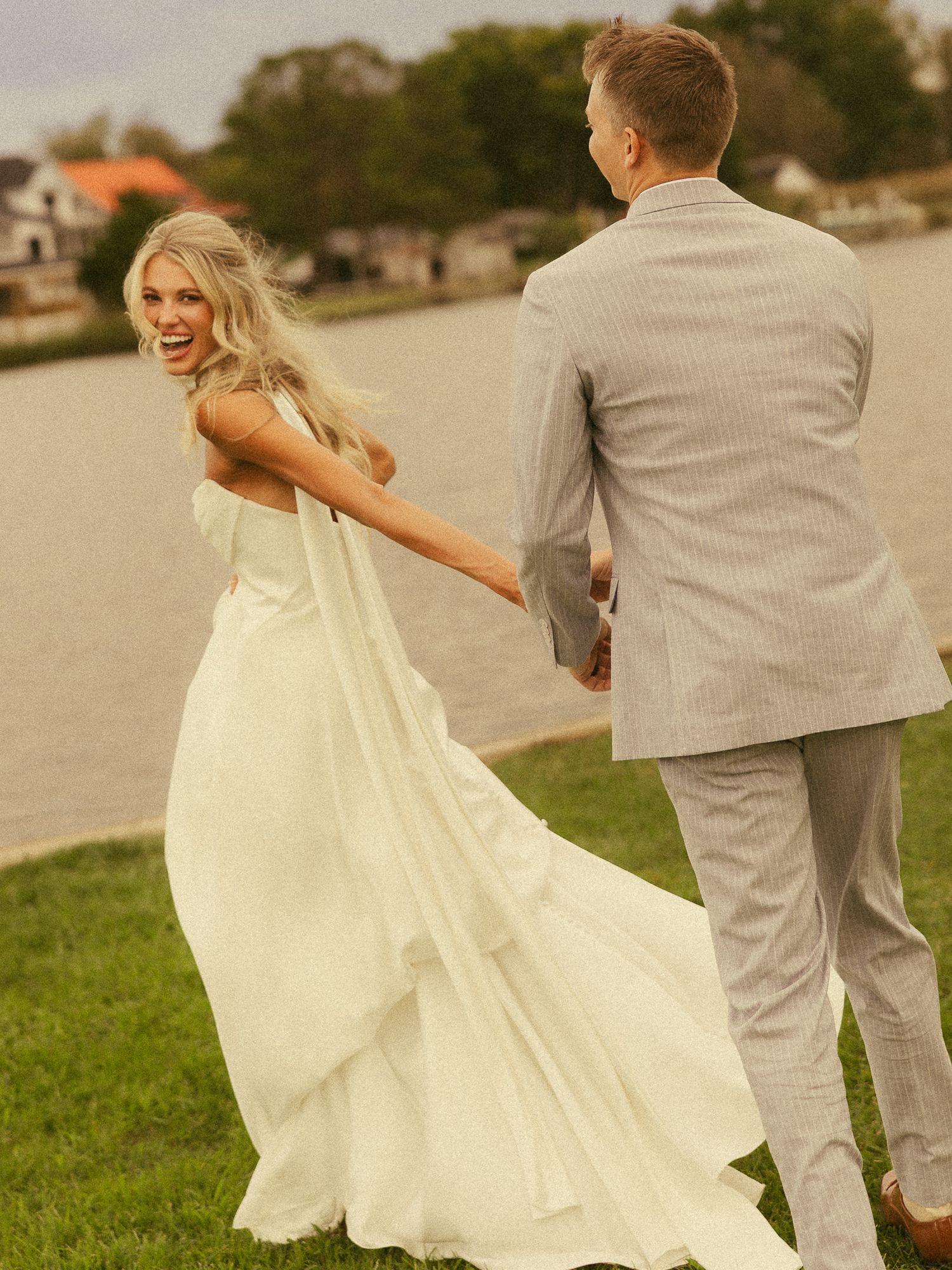 A joyful bride in a white gown holds hands with a groom in a gray suit near a lakeside on their wedding day.