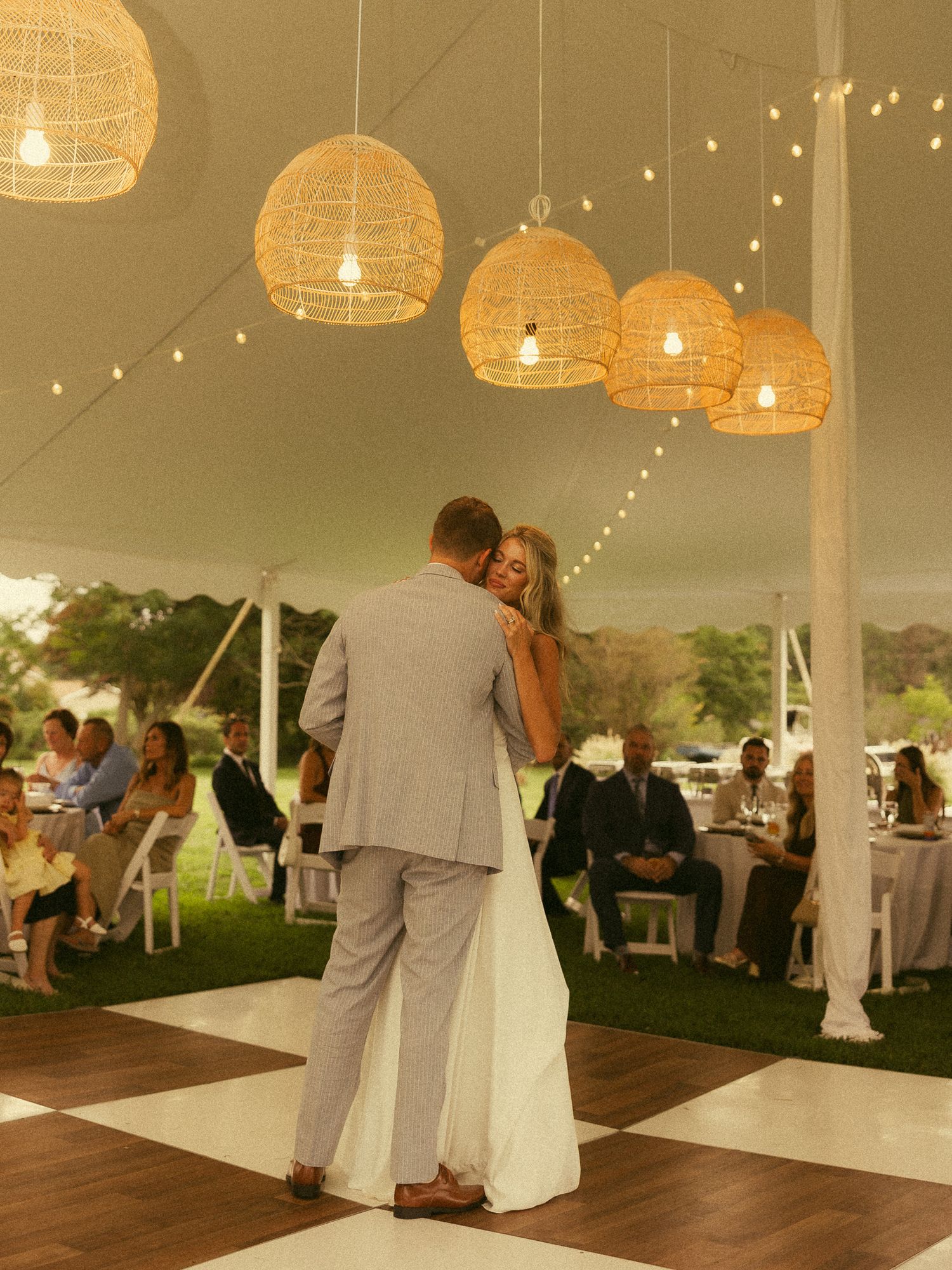 Bride and groom share first dance under wicker pendant lights on checkered dance floor at elegant outdoor tent wedding reception.