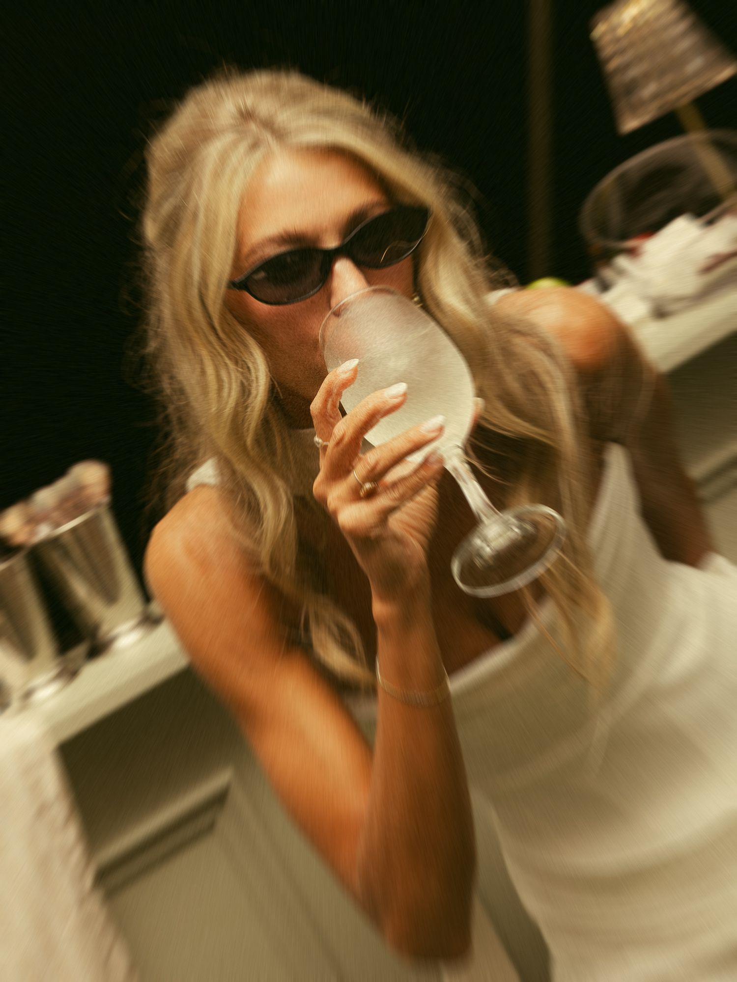 Blonde woman in white dress and black sunglasses sipping from a large wine glass at an elegant dinner event.