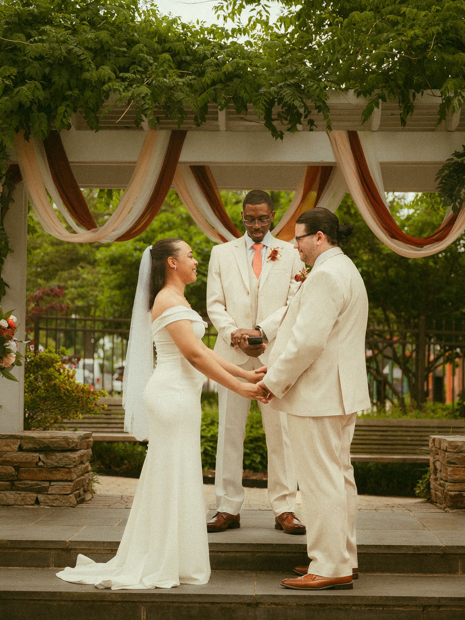 Bride and groom hold hands at outdoor wedding ceremony under floral pergola with officiant.