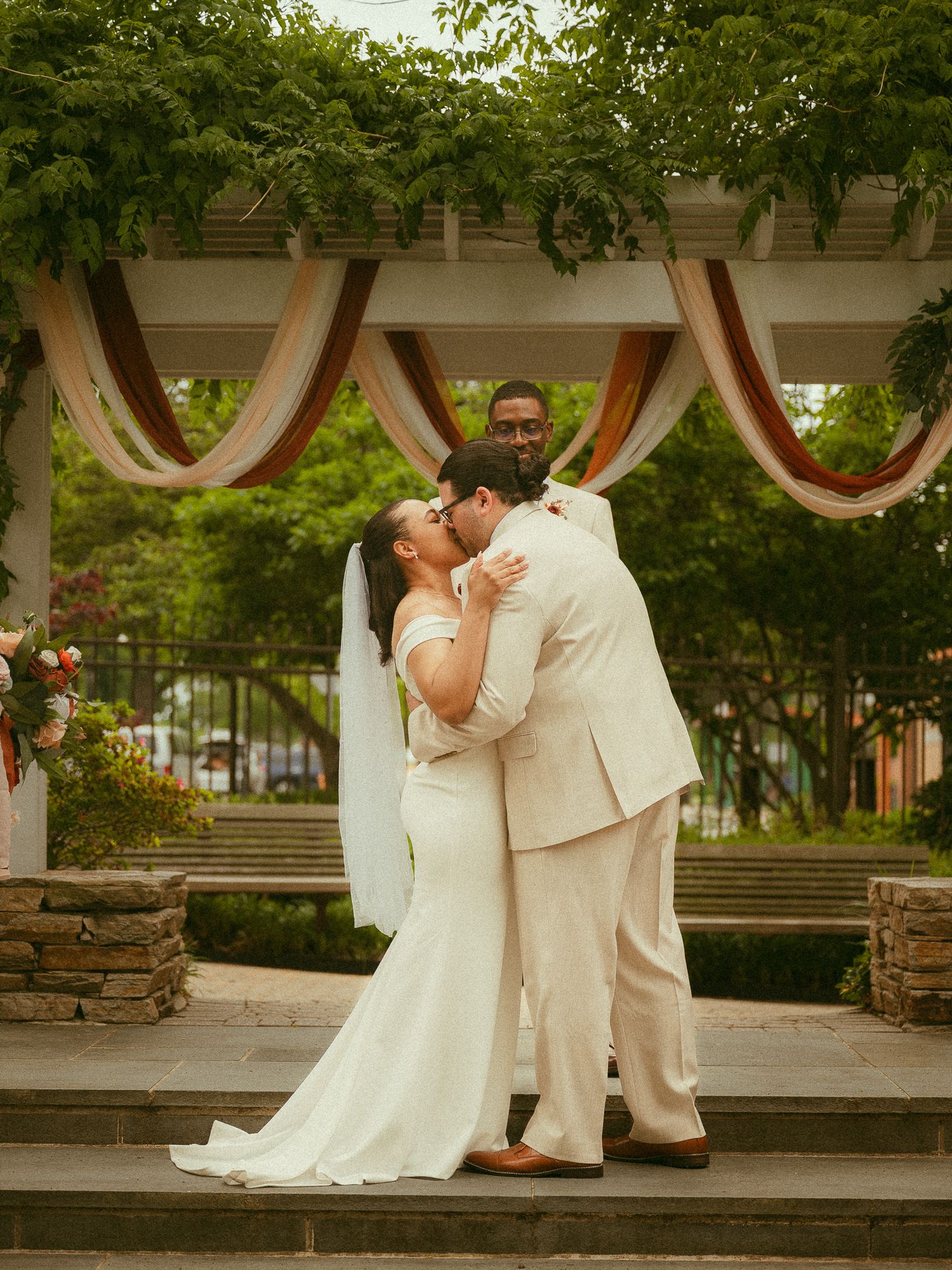 Newlyweds share first kiss under floral pergola draped with burgundy and cream fabric at outdoor wedding.