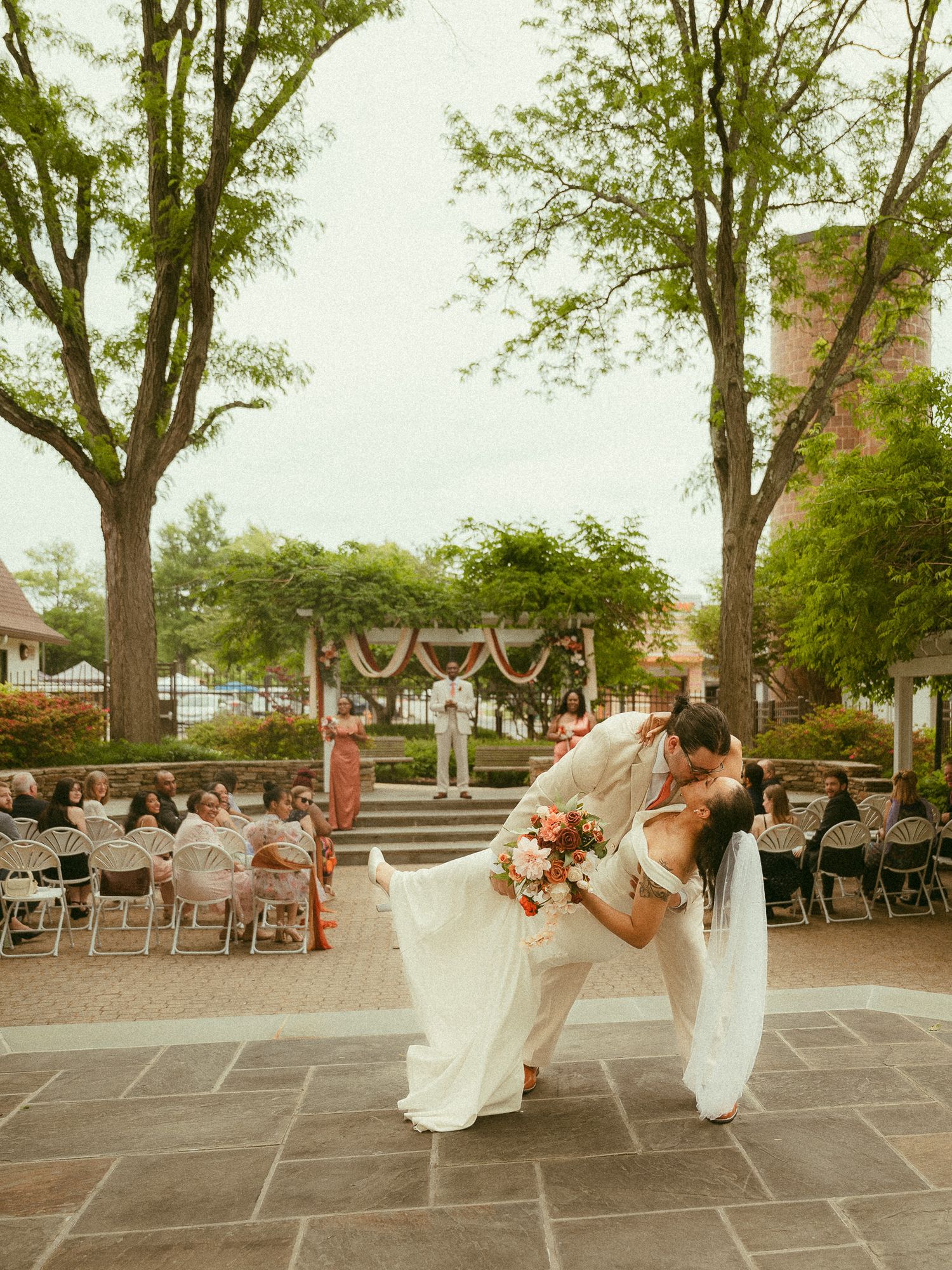 Bride and groom share a romantic dip kiss during outdoor wedding ceremony with guests seated on patio.