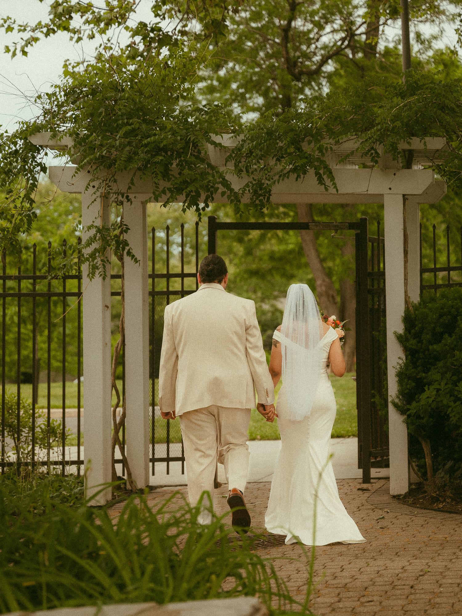 Bride and groom walking hand in hand through a white garden arbor gate surrounded by lush greenery.