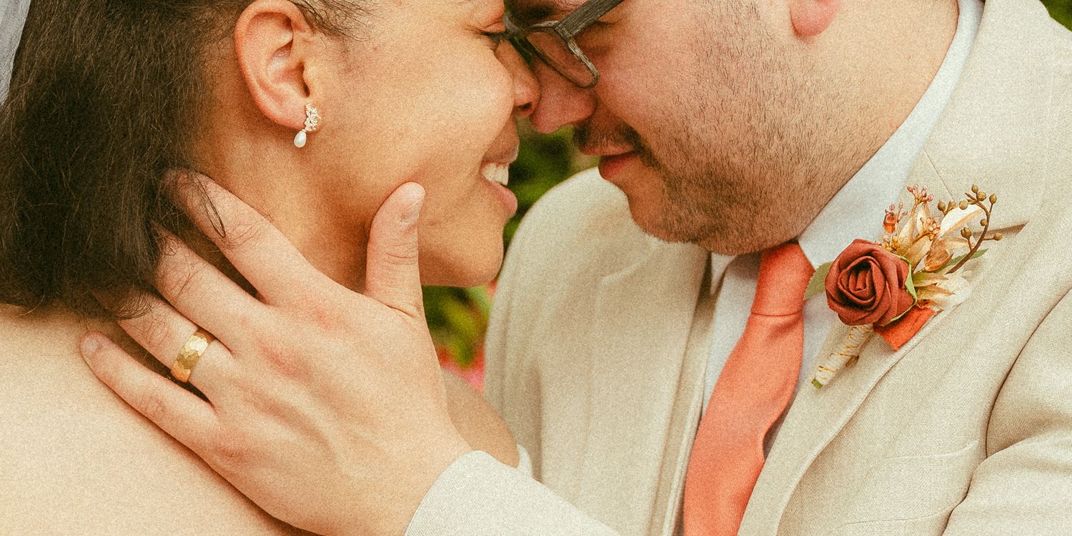 A joyful couple in wedding attire shares an intimate moment, the groom in a tan suit with coral tie and rose boutonniere.