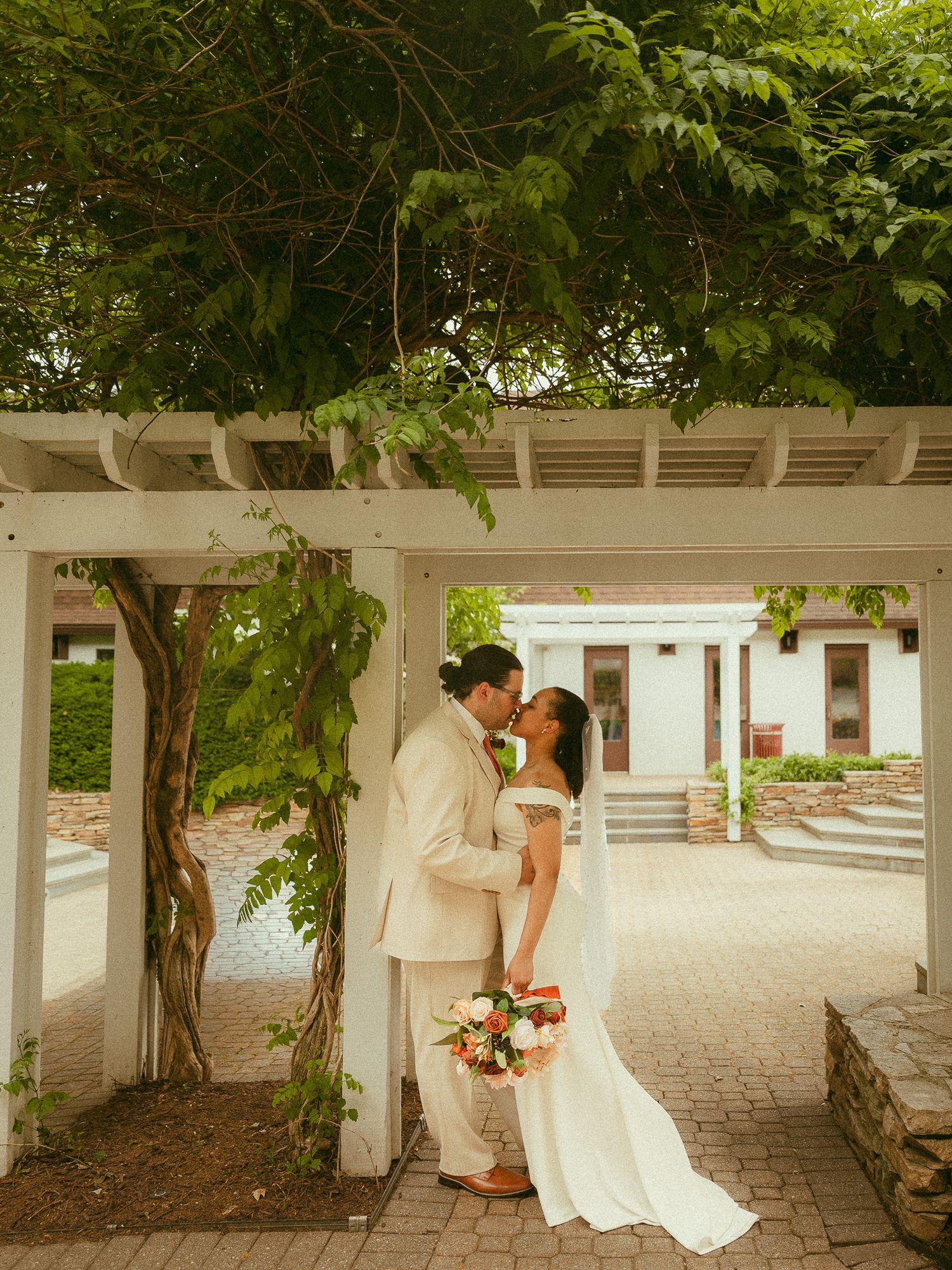 A bride and groom share a kiss under a white pergola covered in lush greenery, bride holding a bouquet.