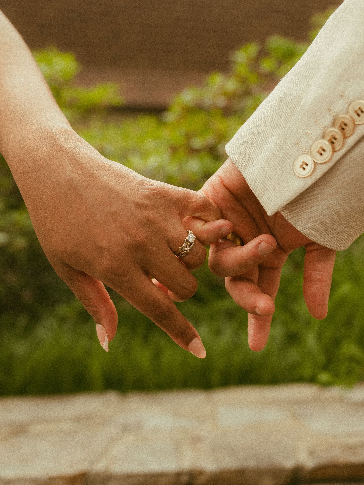Close-up of newlyweds holding hands showing engagement ring and wedding band outdoors.