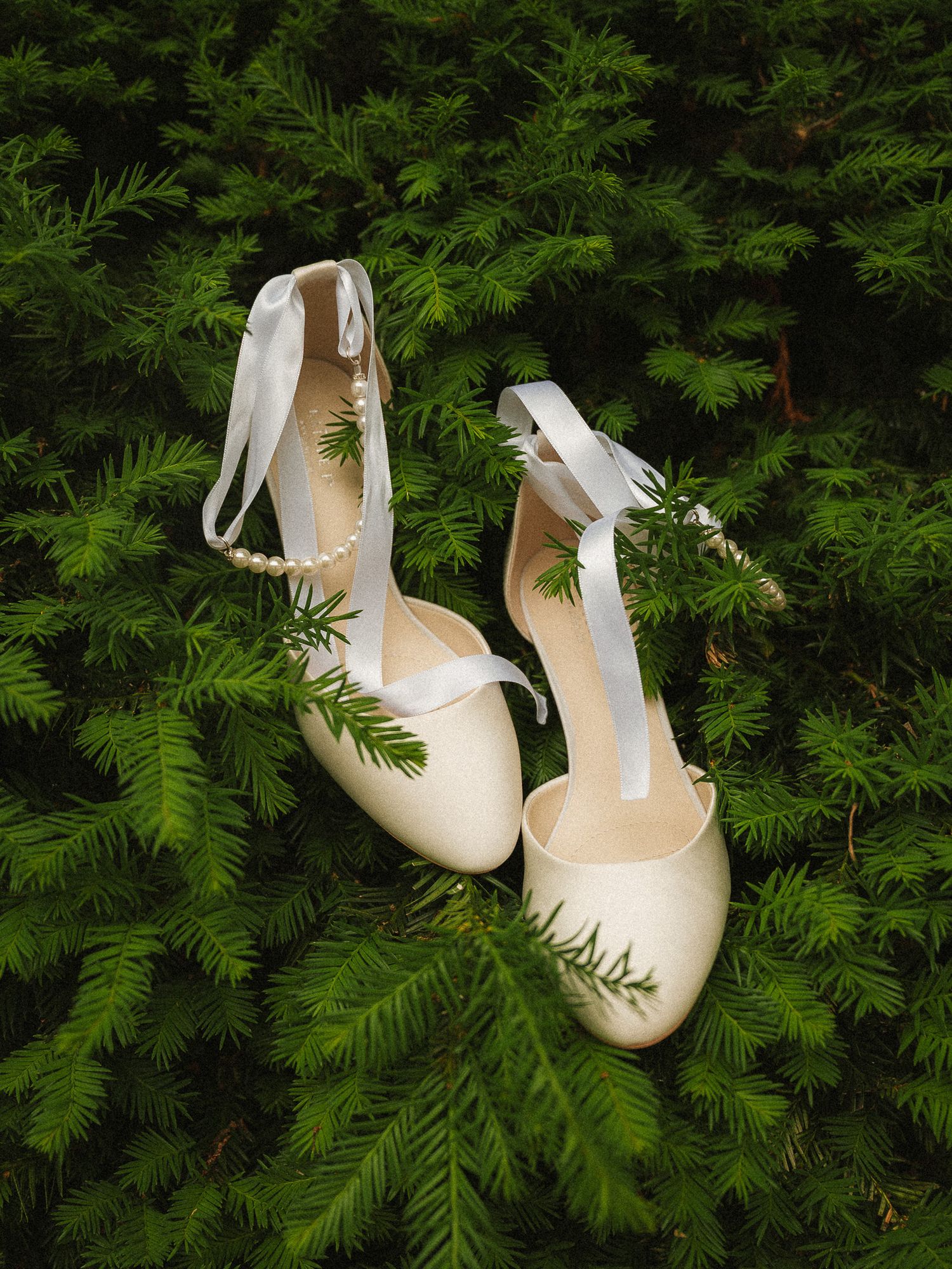 Elegant ivory bridal heels with satin ribbon ankle ties and pearl accents resting on lush green pine branches.