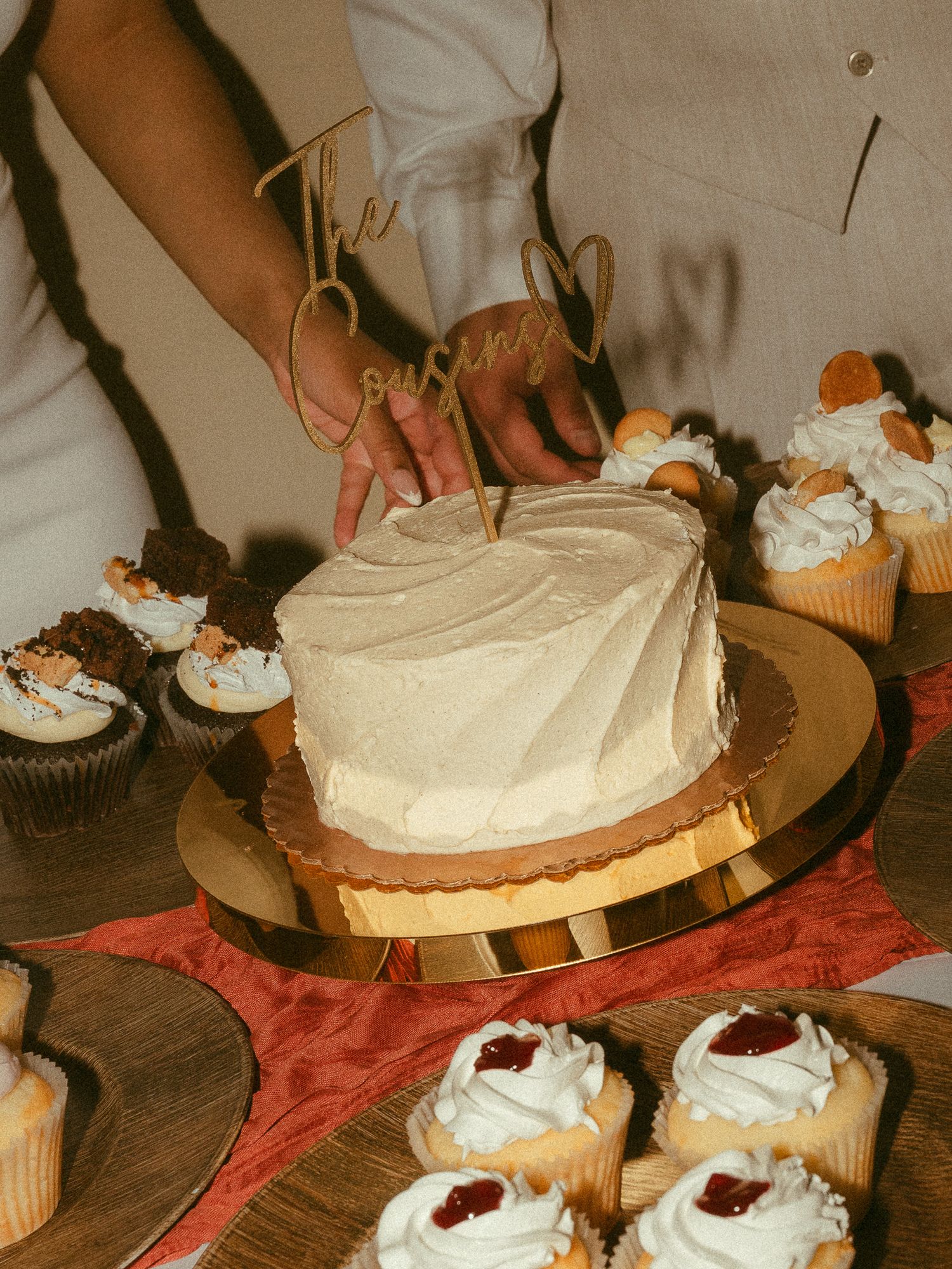 Hands cutting a white frosted wedding cake with a gold topper surrounded by assorted cupcakes on a table.