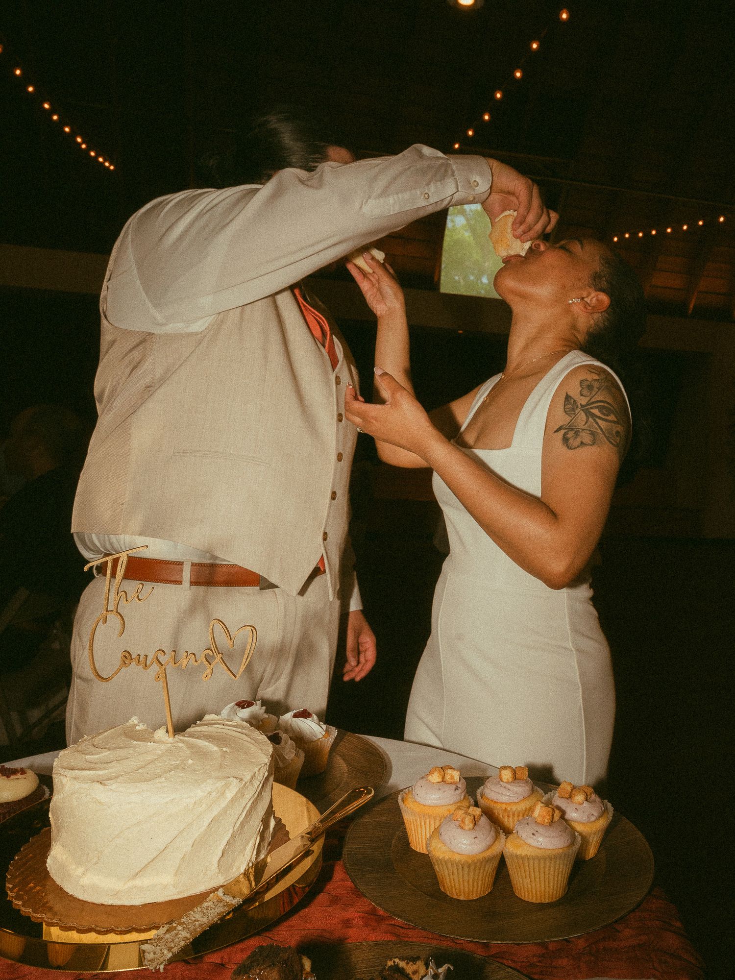 Couple feeds each other wedding cake at reception with white frosted cake and cupcakes on display.