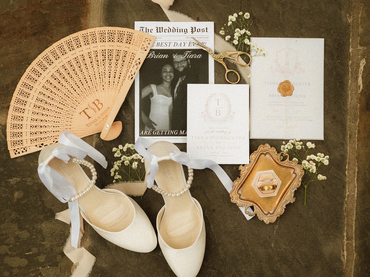 Elegant wedding flat lay with white heels, pearl details, invitation suite, wooden fan, and floral accents on stone surface.