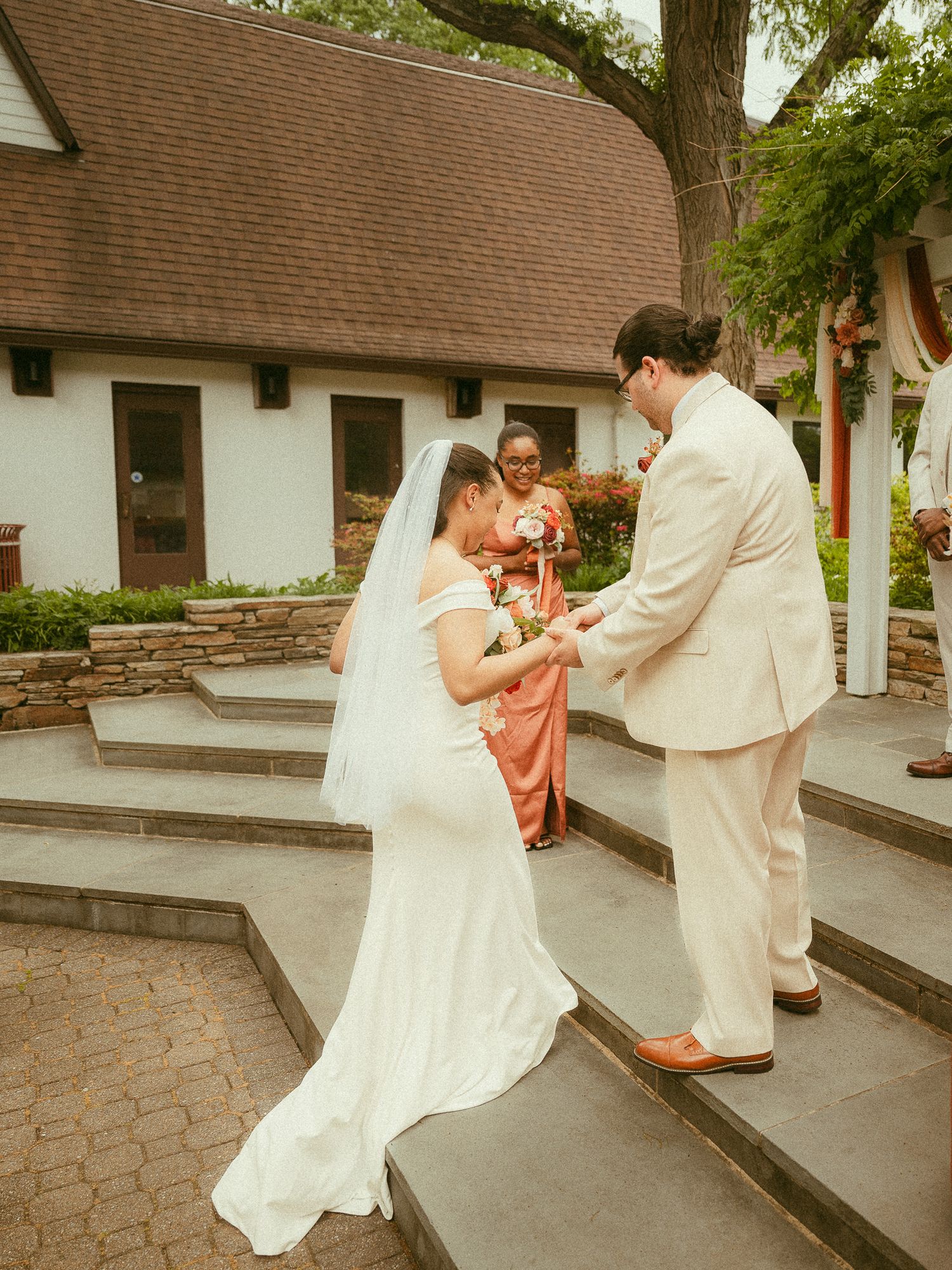 Bride and groom exchange rings on stone steps outdoors with bridesmaid in coral dress during wedding ceremony.