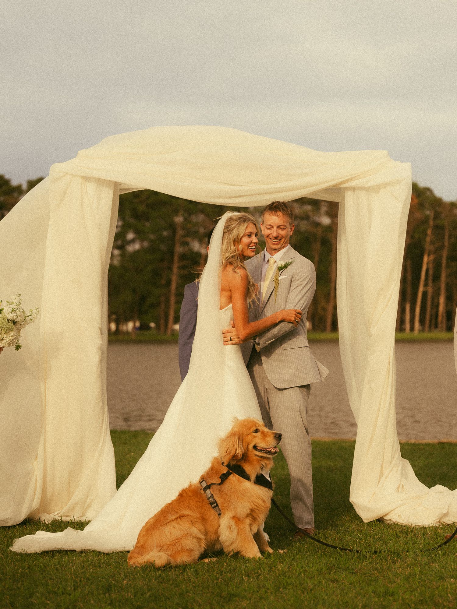 Bride and groom pose under white draped arch with golden retriever dog at outdoor waterfront wedding ceremony.