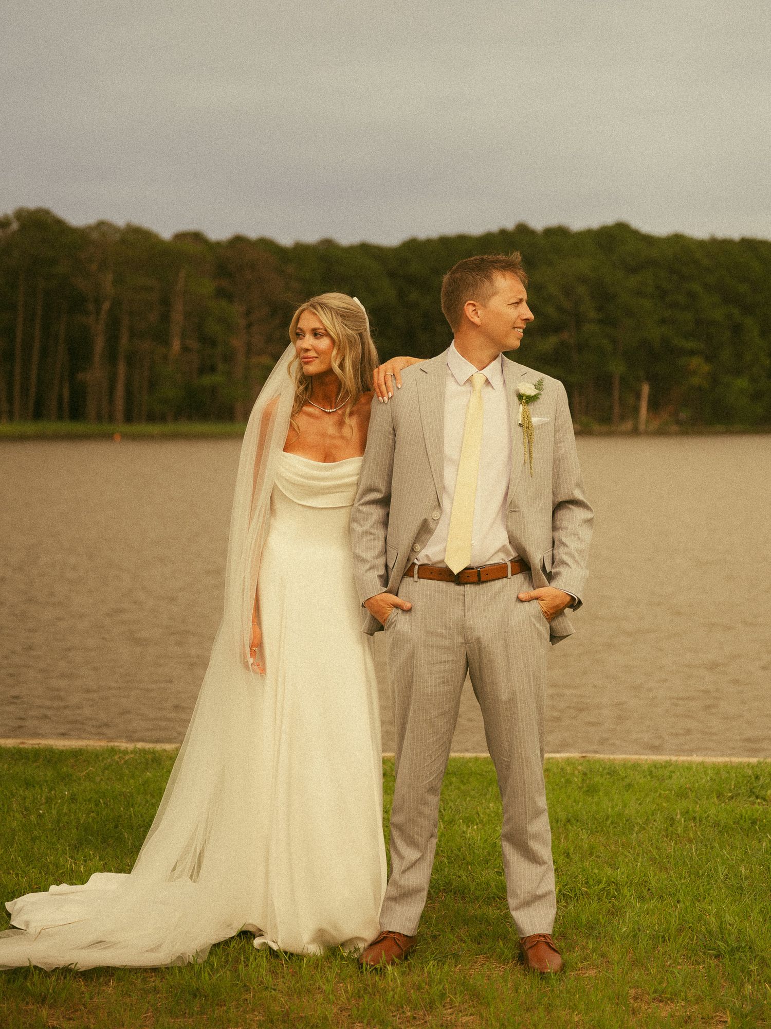 Bride in white gown with veil and groom in gray suit pose by a serene lake surrounded by lush trees at golden hour.