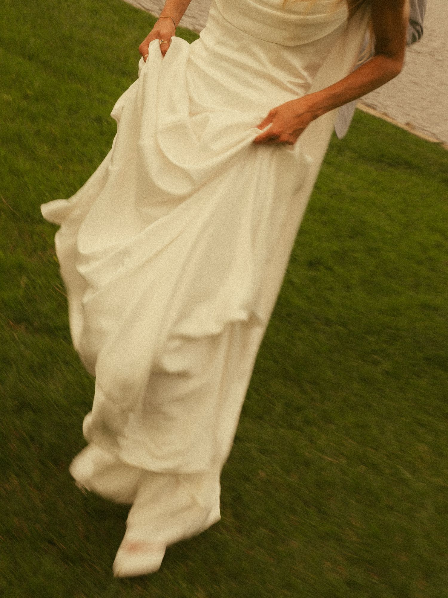 Bride in flowing white wedding gown holding dress hem while walking on lush green grass outdoors.