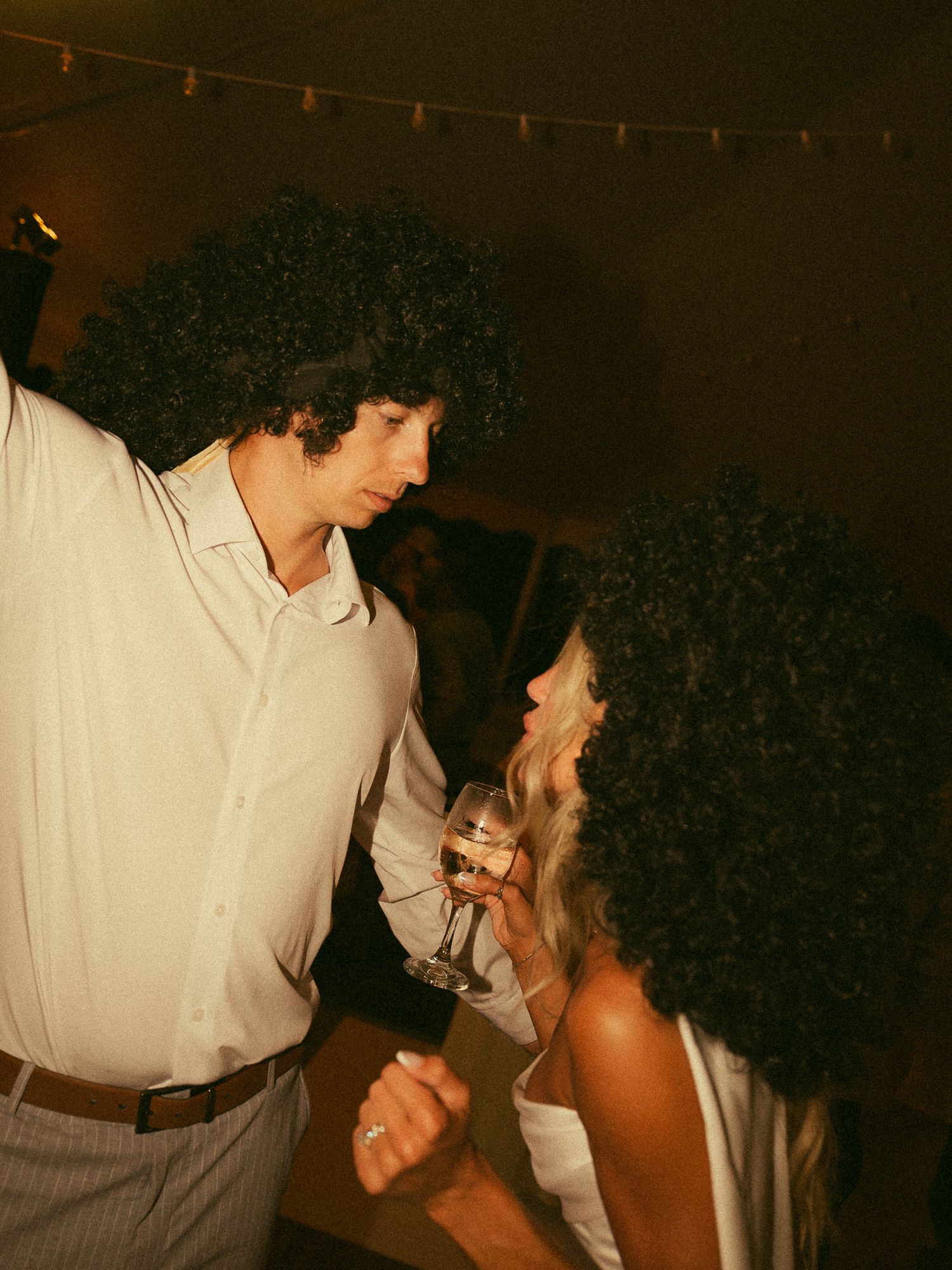 Two people wearing curly afro wigs dancing and socializing at a party with string lights in background.