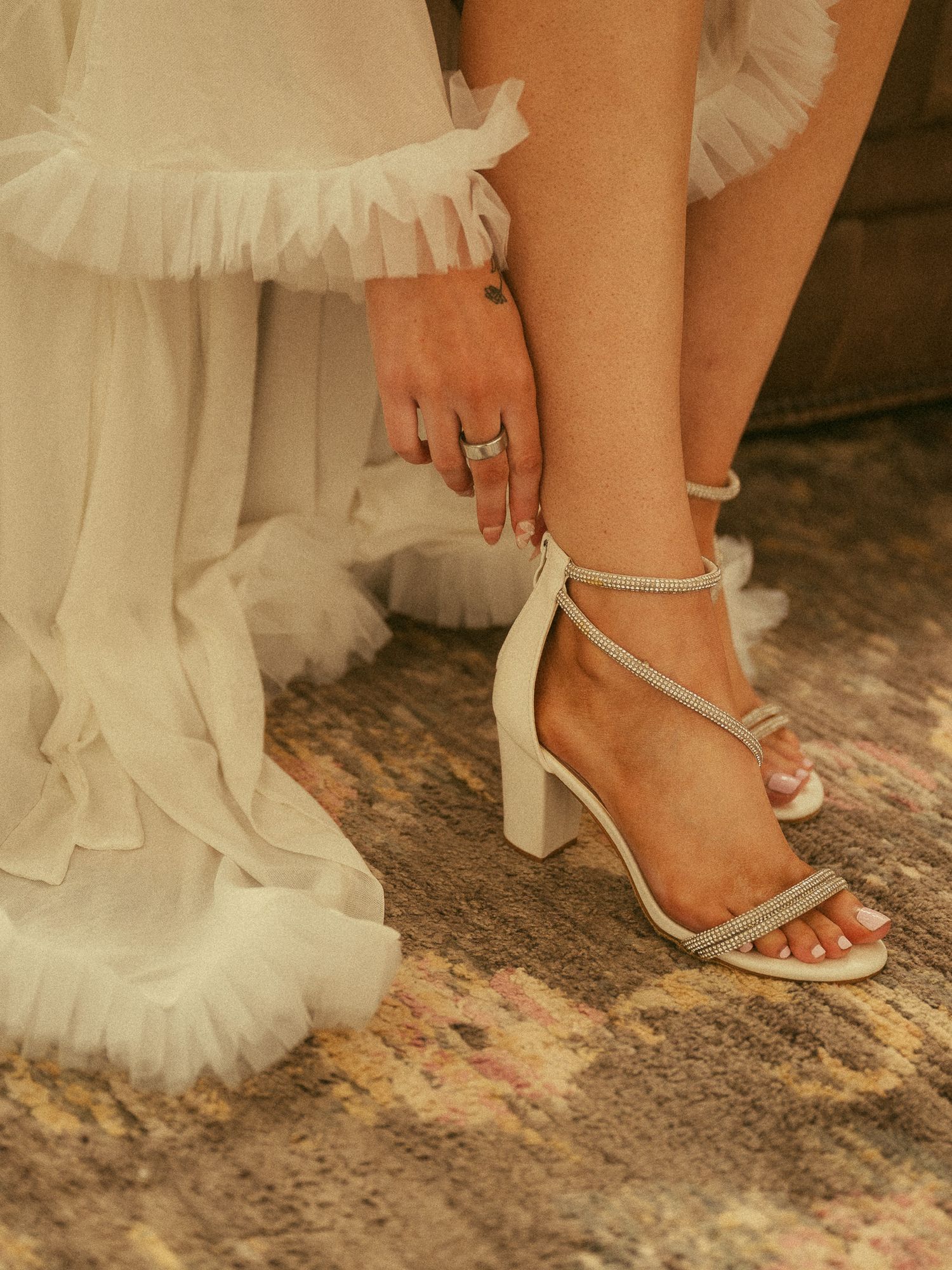 Bride wearing white block heel sandals with crystal straps, adjusting ankle strap while wearing a tulle wedding gown.
