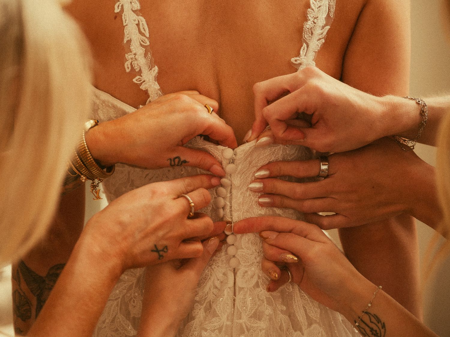 Multiple hands with rings and tattoos helping fasten the back of a white lace wedding dress on a bride.