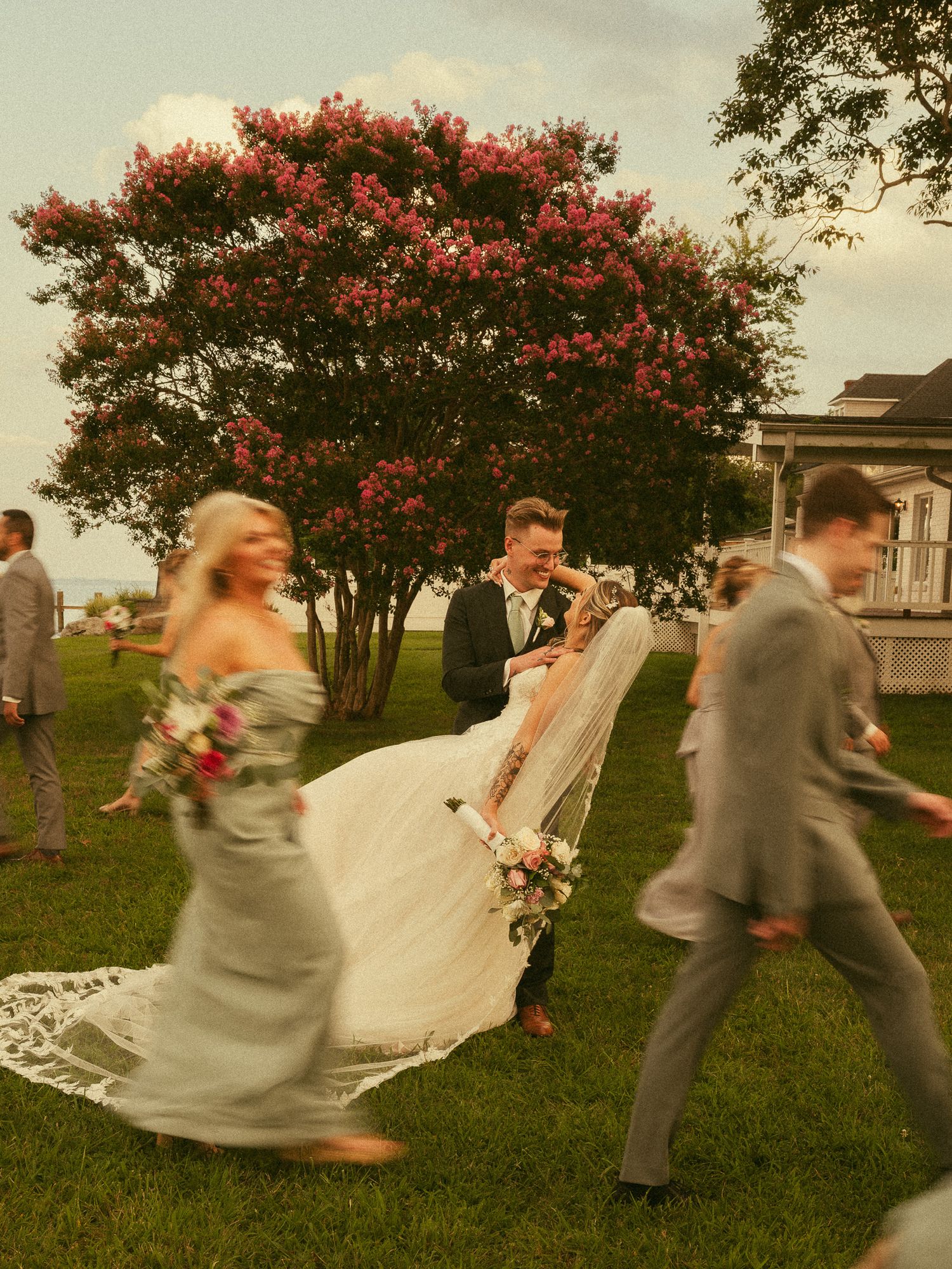 Bride and groom share a kiss surrounded by wedding party on green lawn with blooming pink crepe myrtle tree backdrop.