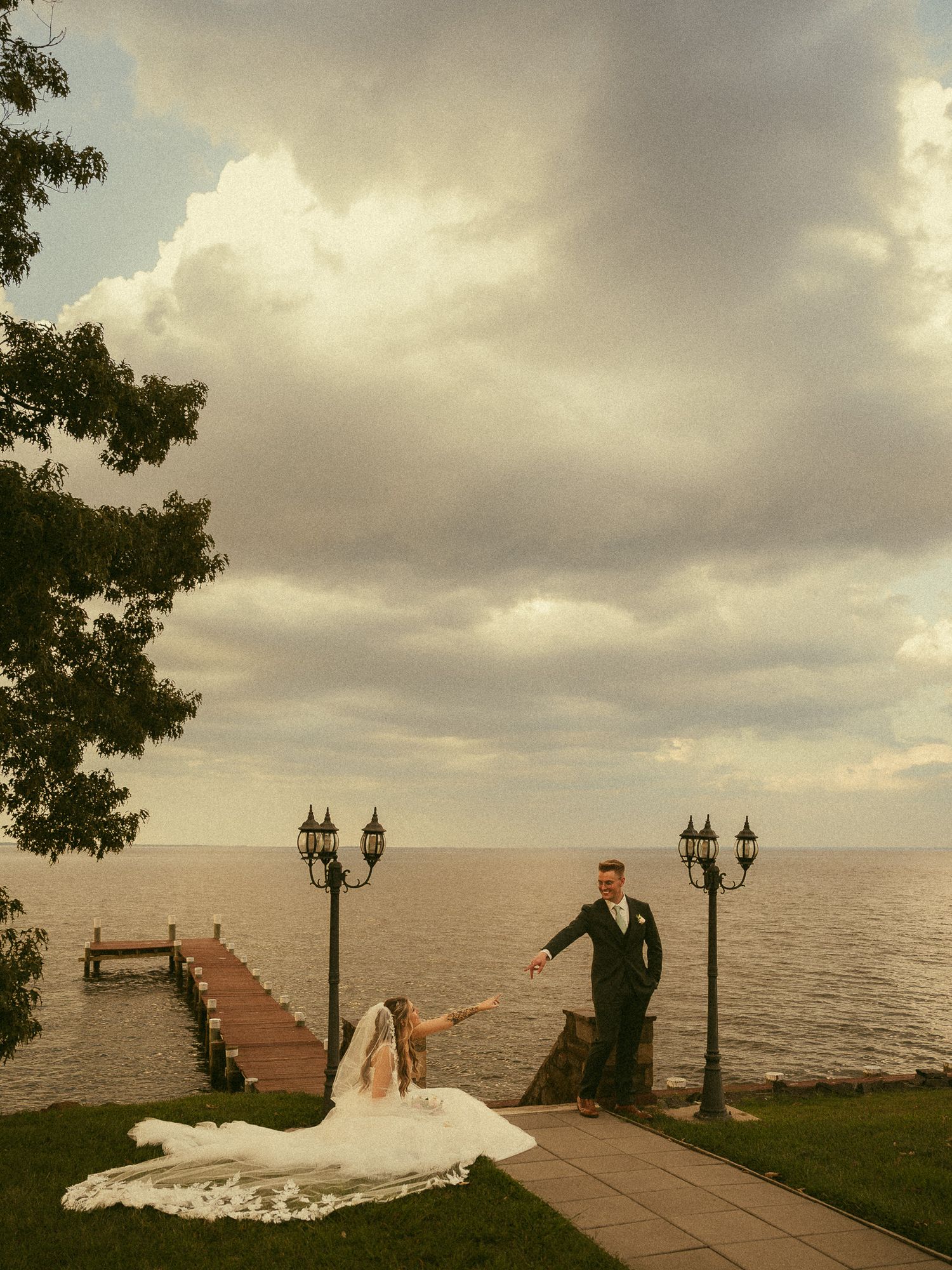 Bride and groom pose by a lakeside dock with dramatic cloudy skies, lamp posts, and a sweeping white wedding gown.