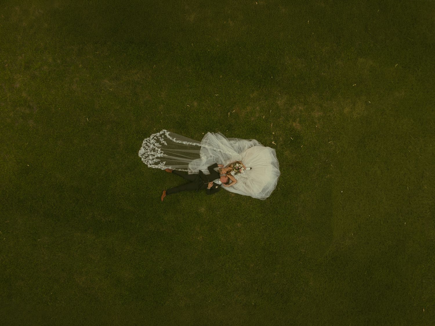 Aerial drone view of a bride in a white wedding dress and veil lying on green grass holding a bouquet.