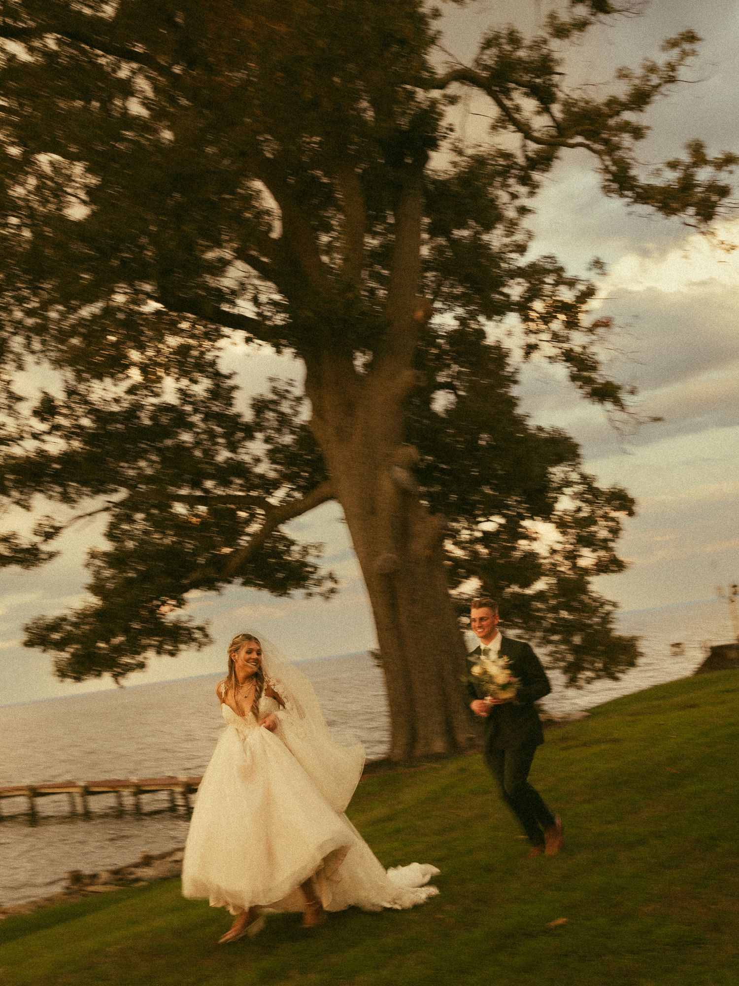 Bride and groom running joyfully on green lawn near large tree by waterfront at golden hour sunset.