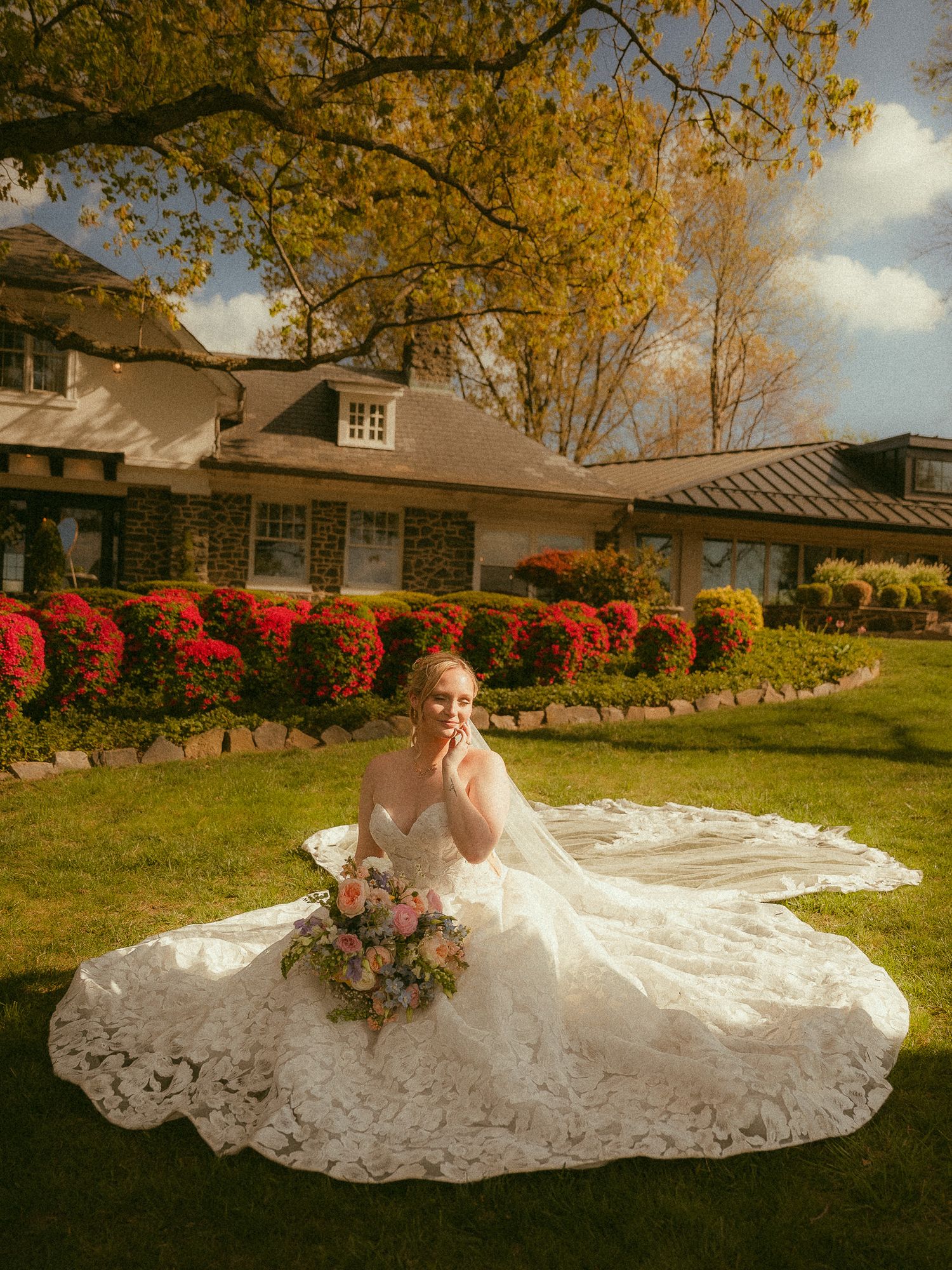 Bride in lace wedding gown sitting on green lawn holding bouquet, with red flowers and cottage in background.