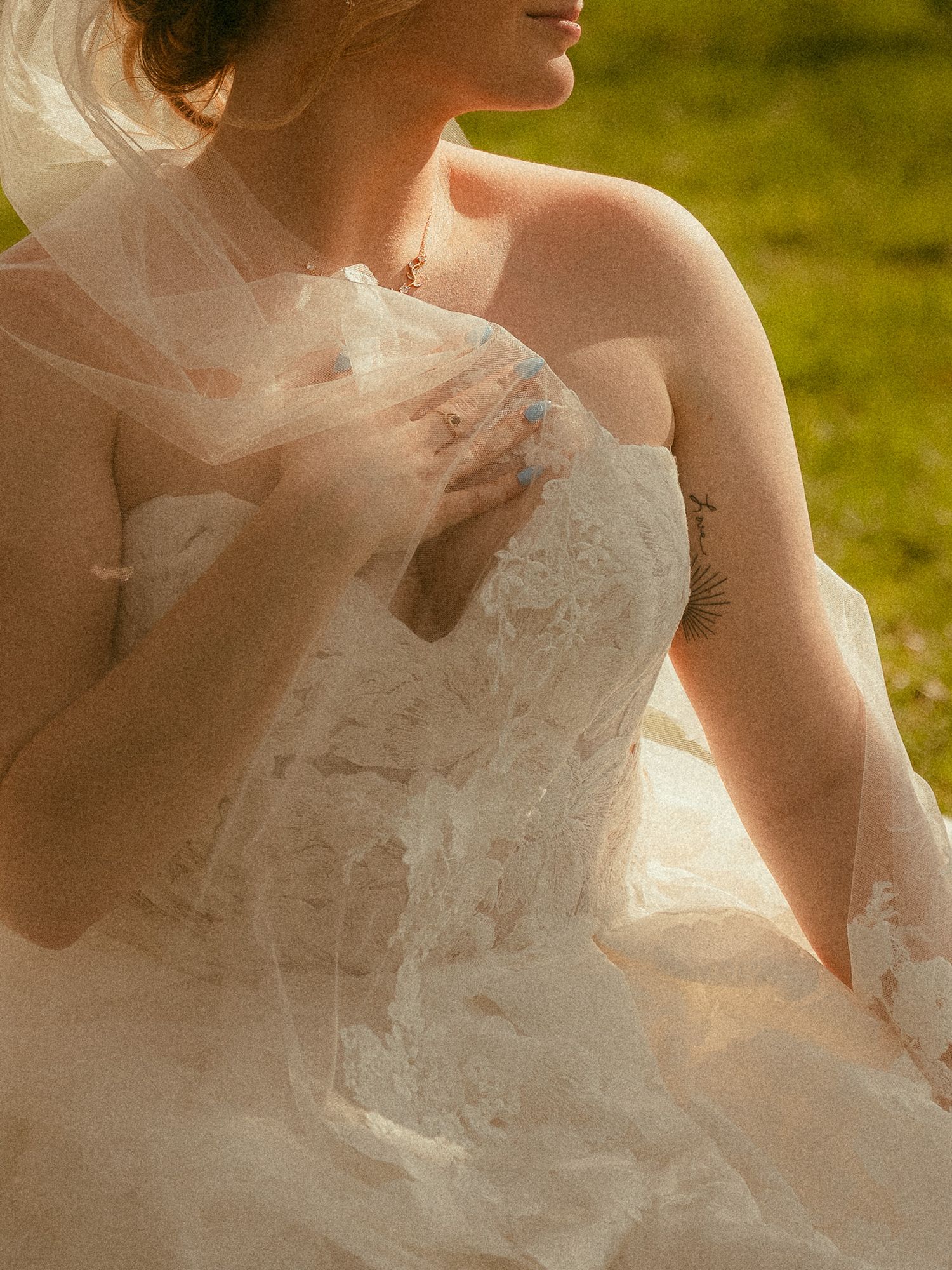 Bride in strapless lace wedding gown with flowing veil seated outdoors in golden sunlight.