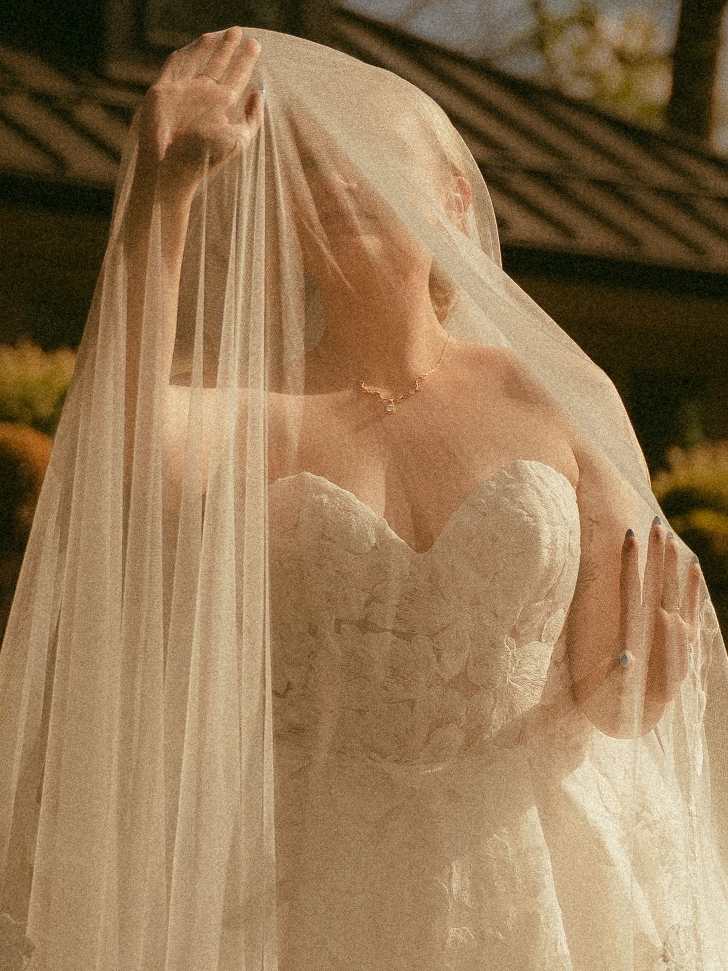 Bride in strapless lace wedding gown lifting sheer veil over face in golden sunlight.