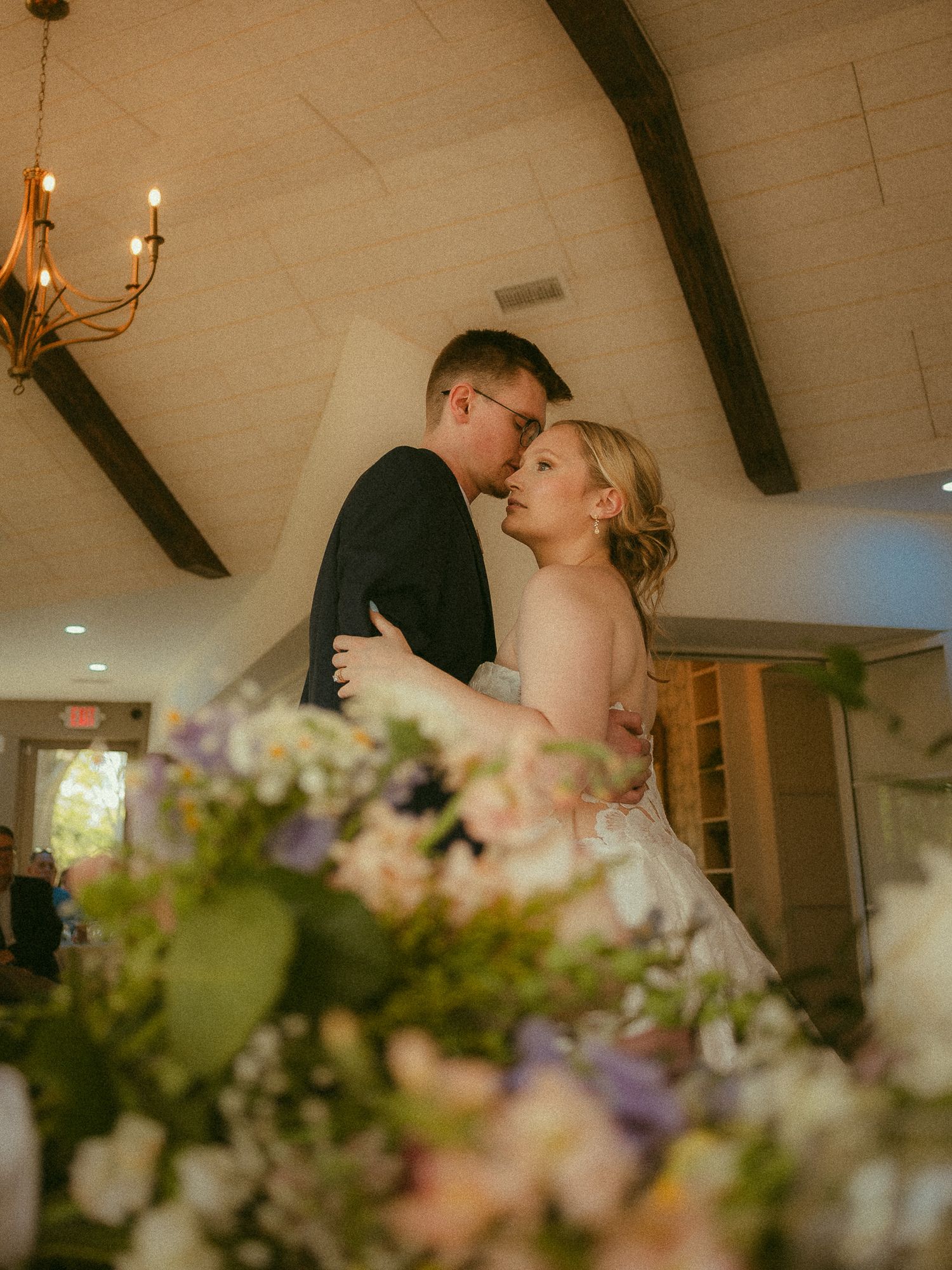 Couple sharing romantic first dance embrace in elegant venue with floral centerpieces and vaulted ceiling.