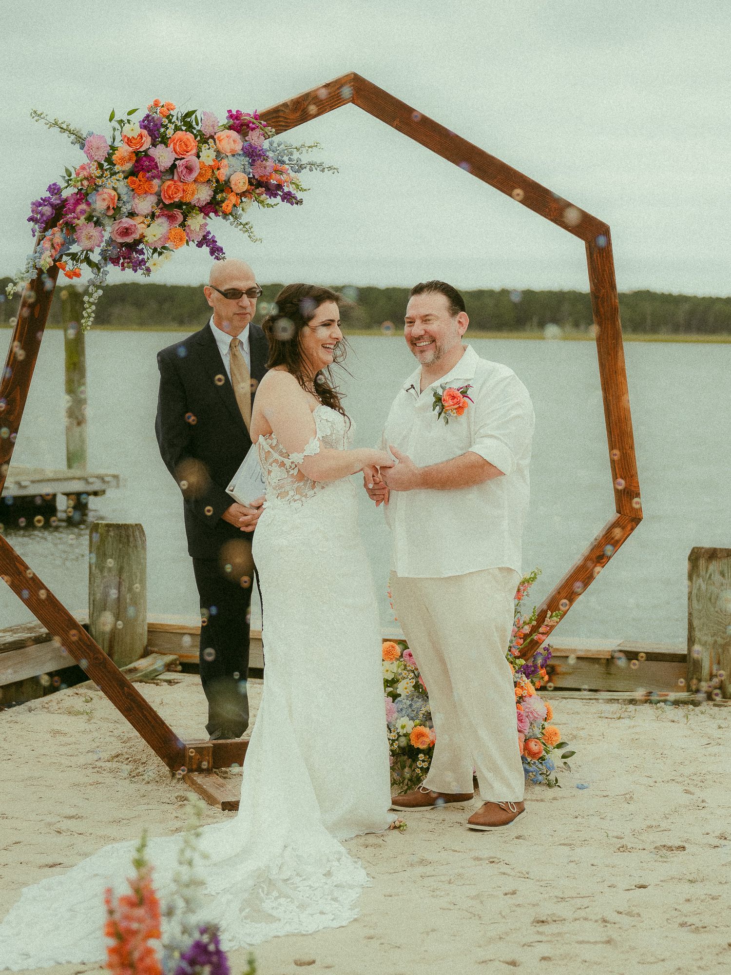 Couple exchanging vows under hexagonal wooden arch with colorful flowers on beach waterfront.