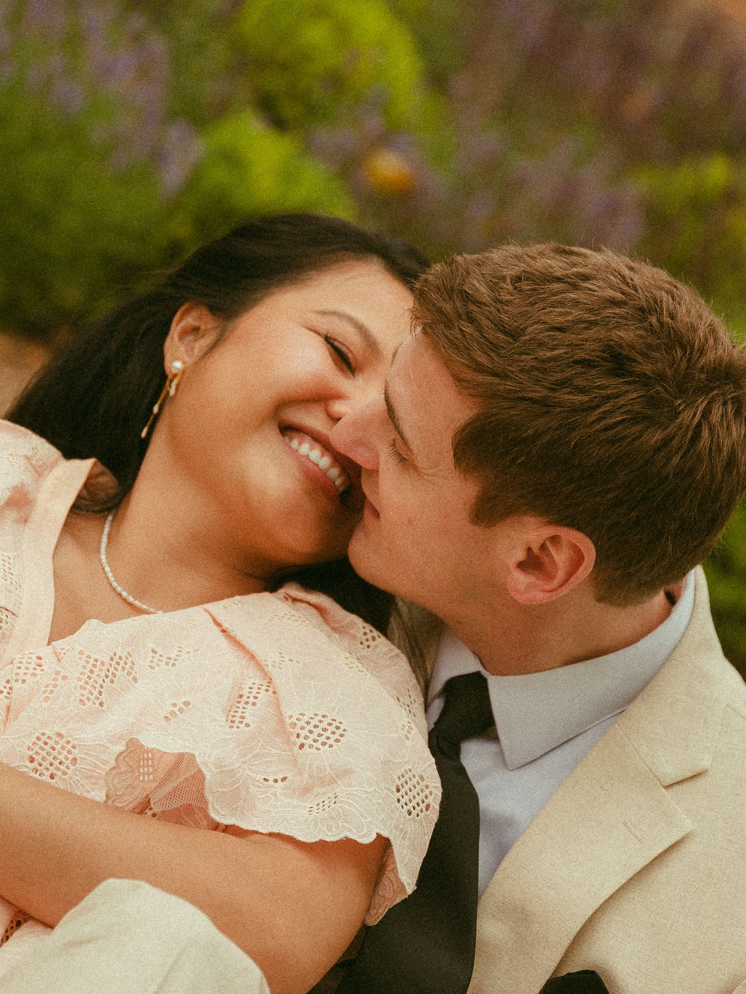 A smiling couple sharing a romantic moment outdoors, with the man nuzzling the laughing woman in a pink dress.