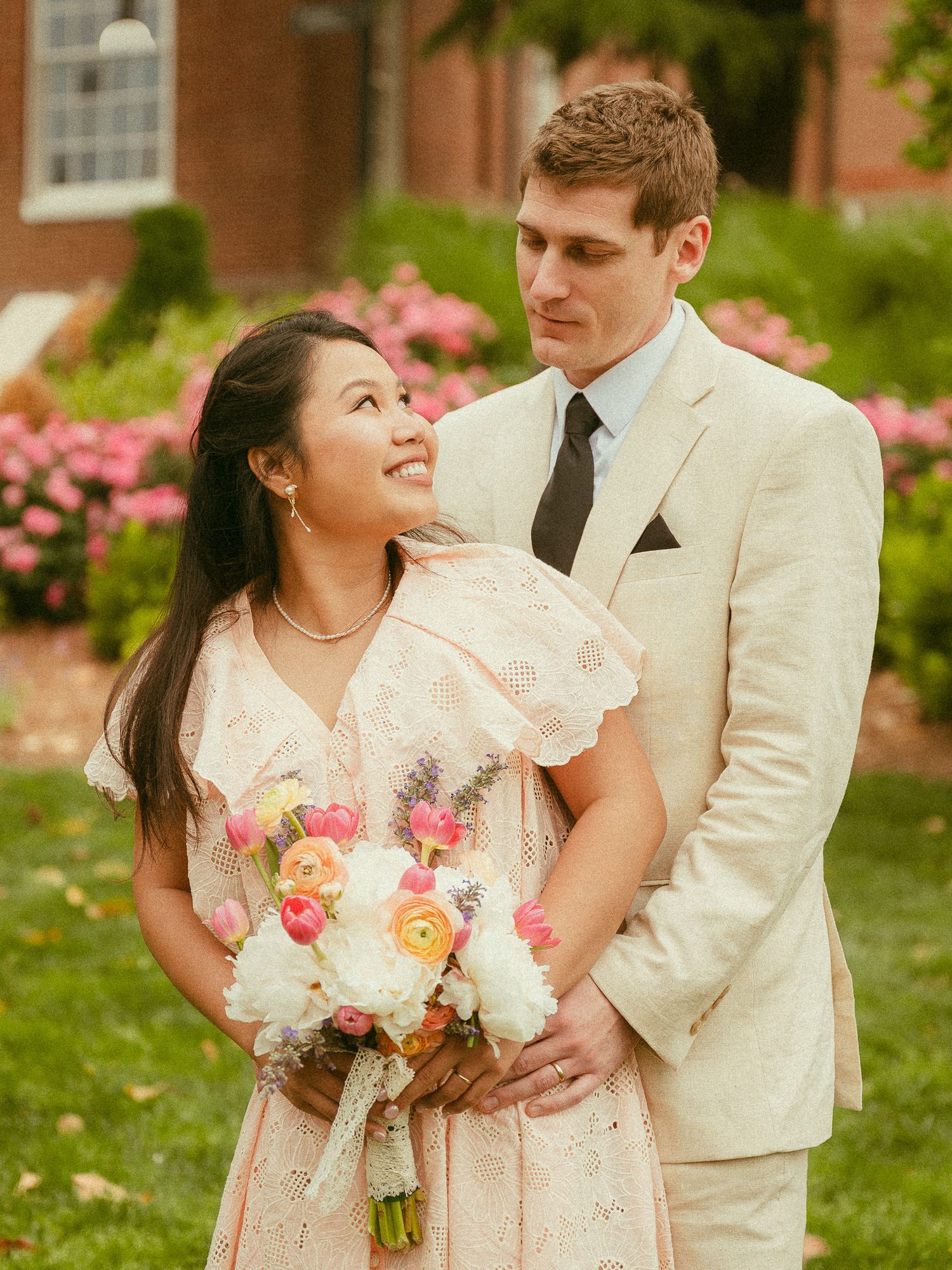 Couple poses outdoors in wedding attire, woman in lace pink dress holding colorful bouquet, man in cream suit.