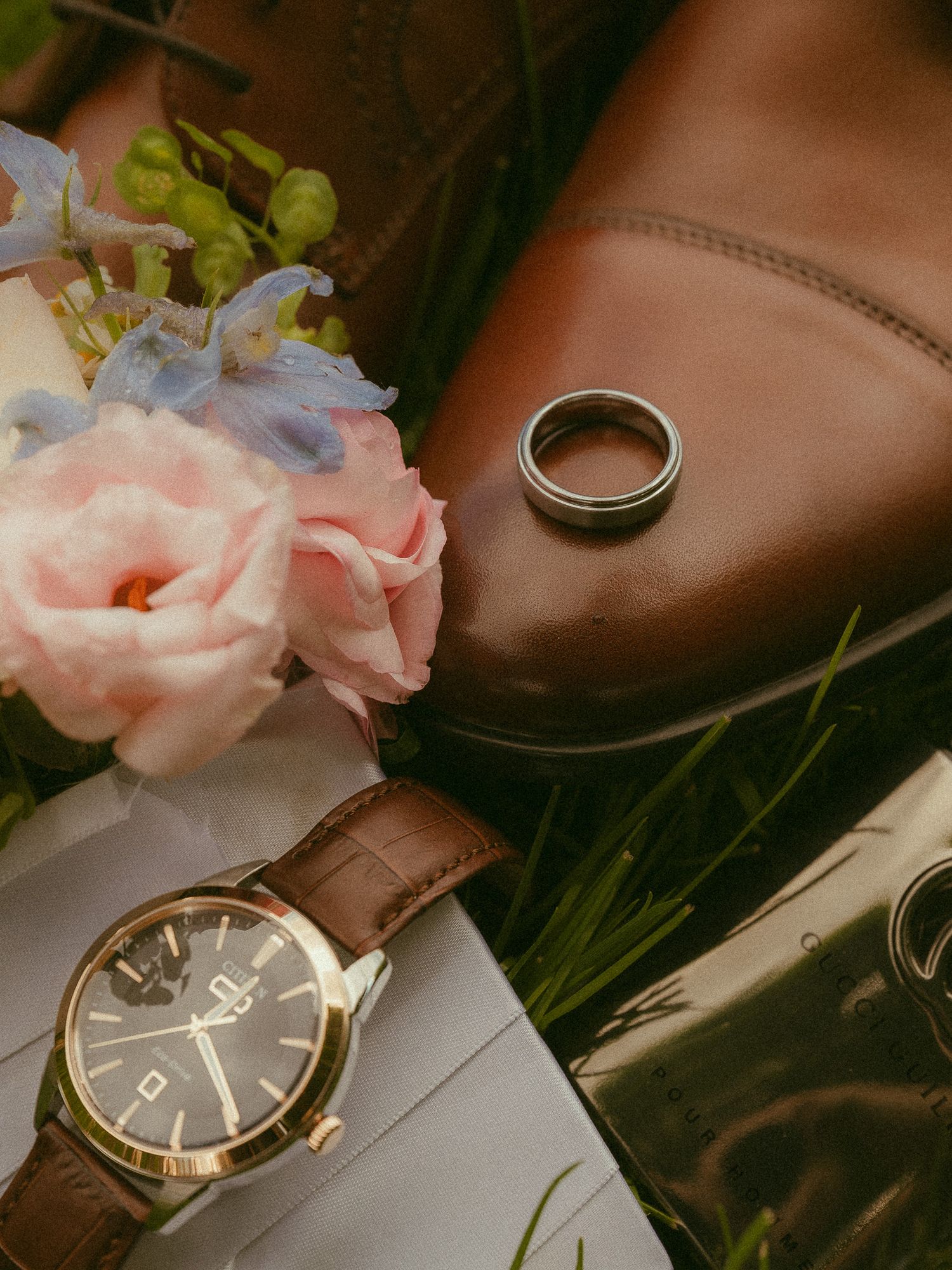 Brown leather shoes, wedding ring, elegant watch with brown strap, and pink and blue flowers arranged for a wedding flat lay.