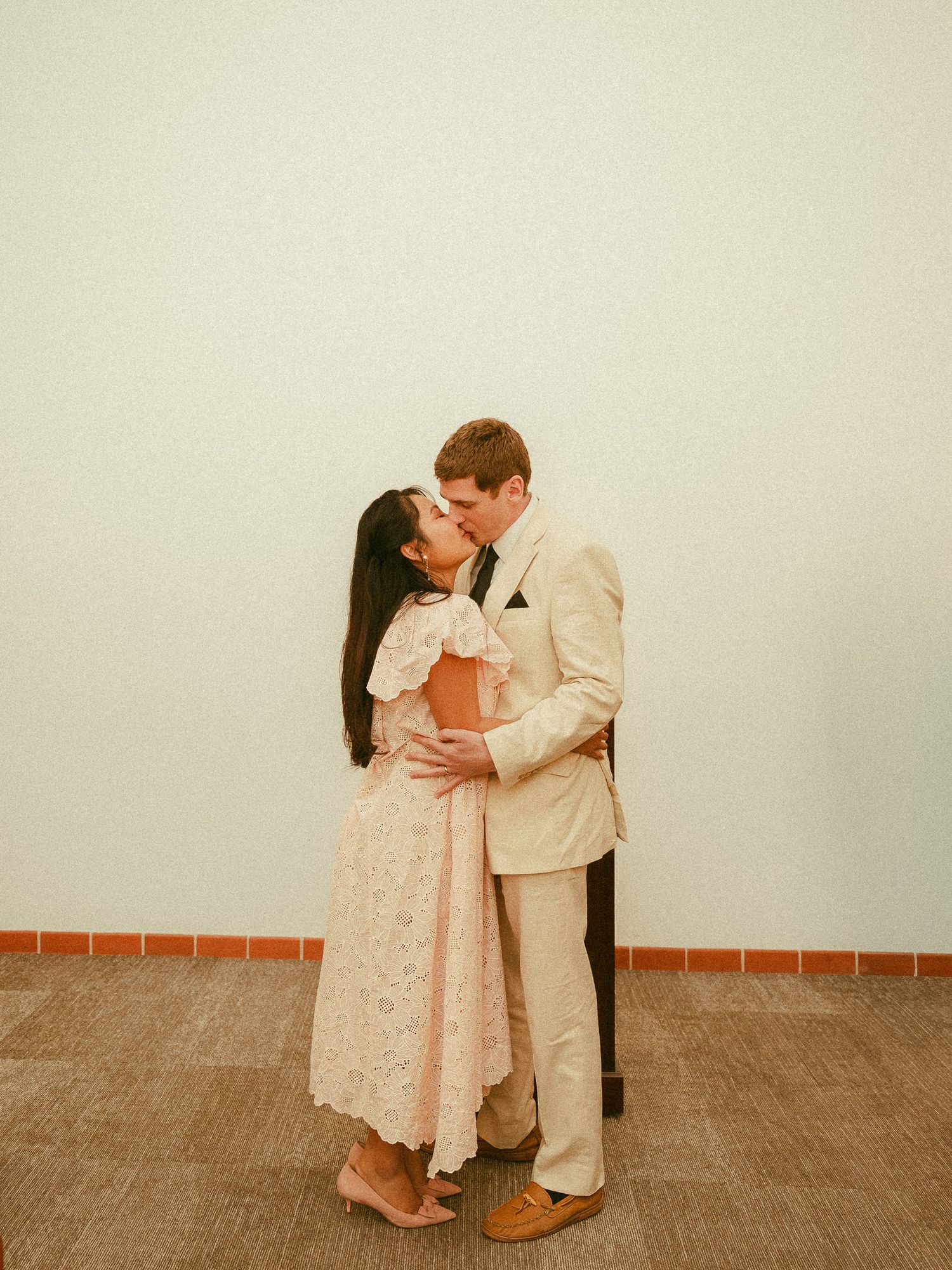 Couple shares a kiss indoors, woman in pink lace dress and man in cream suit embracing tenderly.