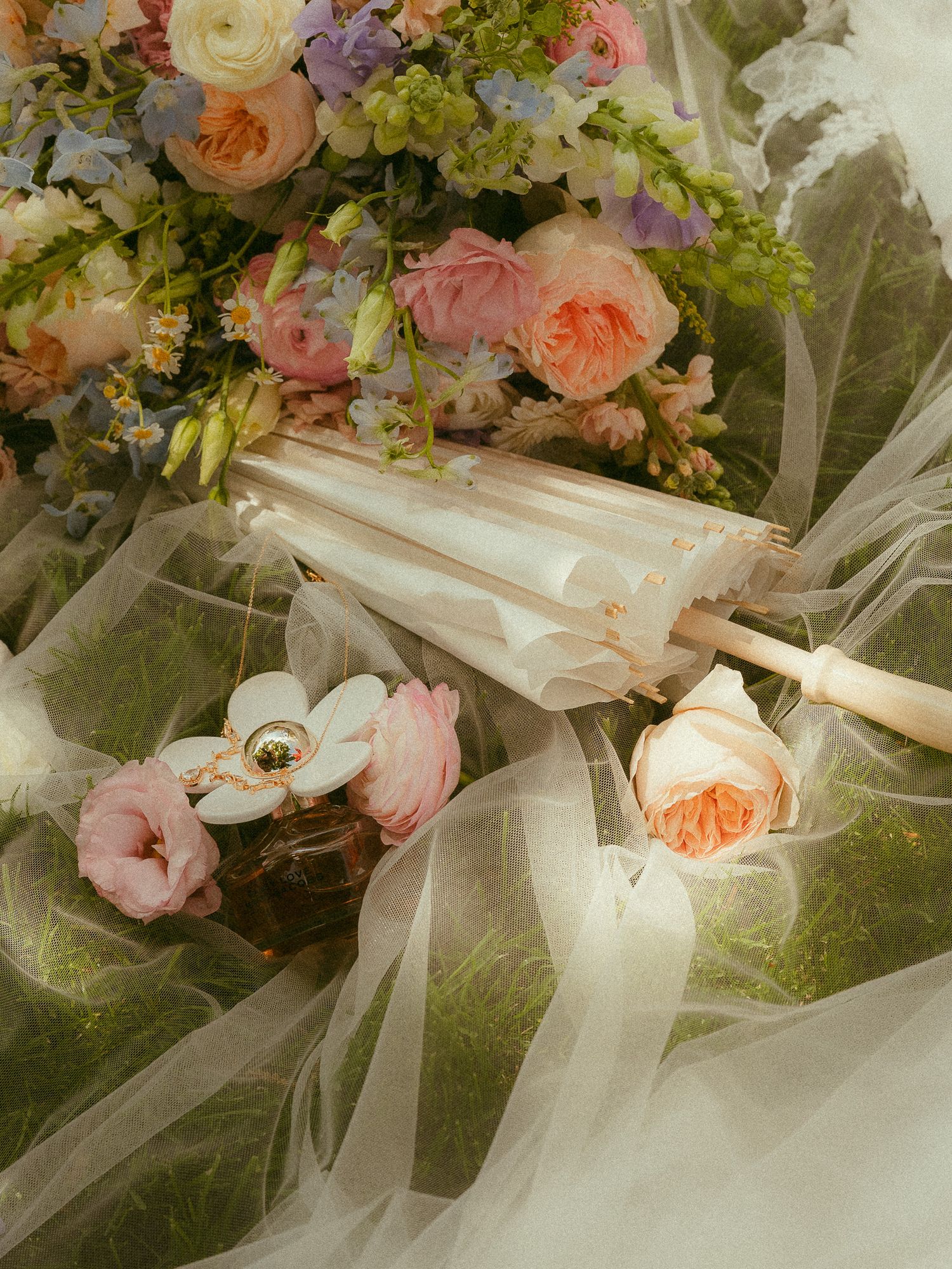 Elegant white parasol with pearl handle resting on sheer tulle beside colorful spring floral bouquet and pink roses.