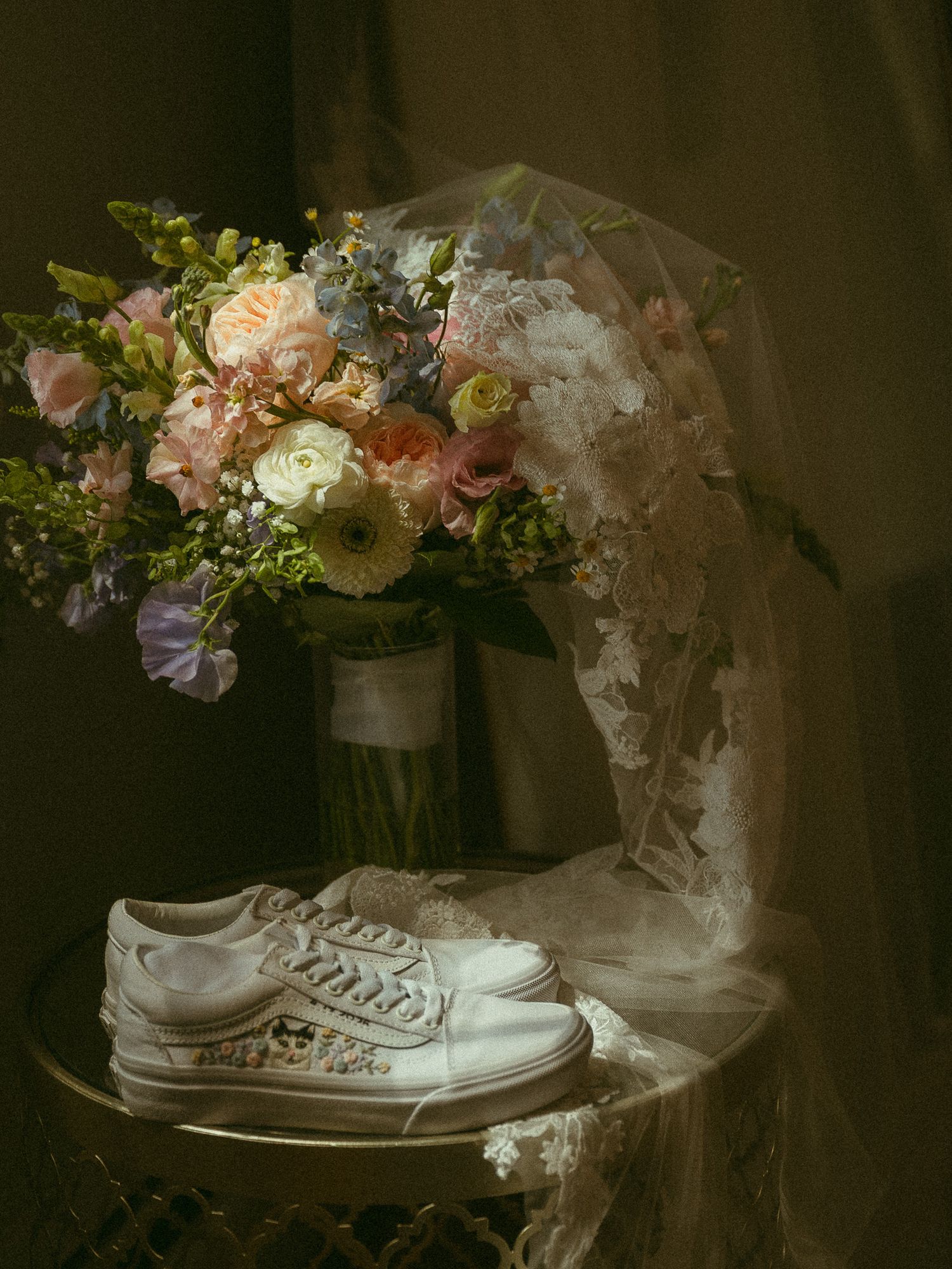 White bridal sneakers rest beside a romantic pastel wedding bouquet and lace veil on a draped table.
