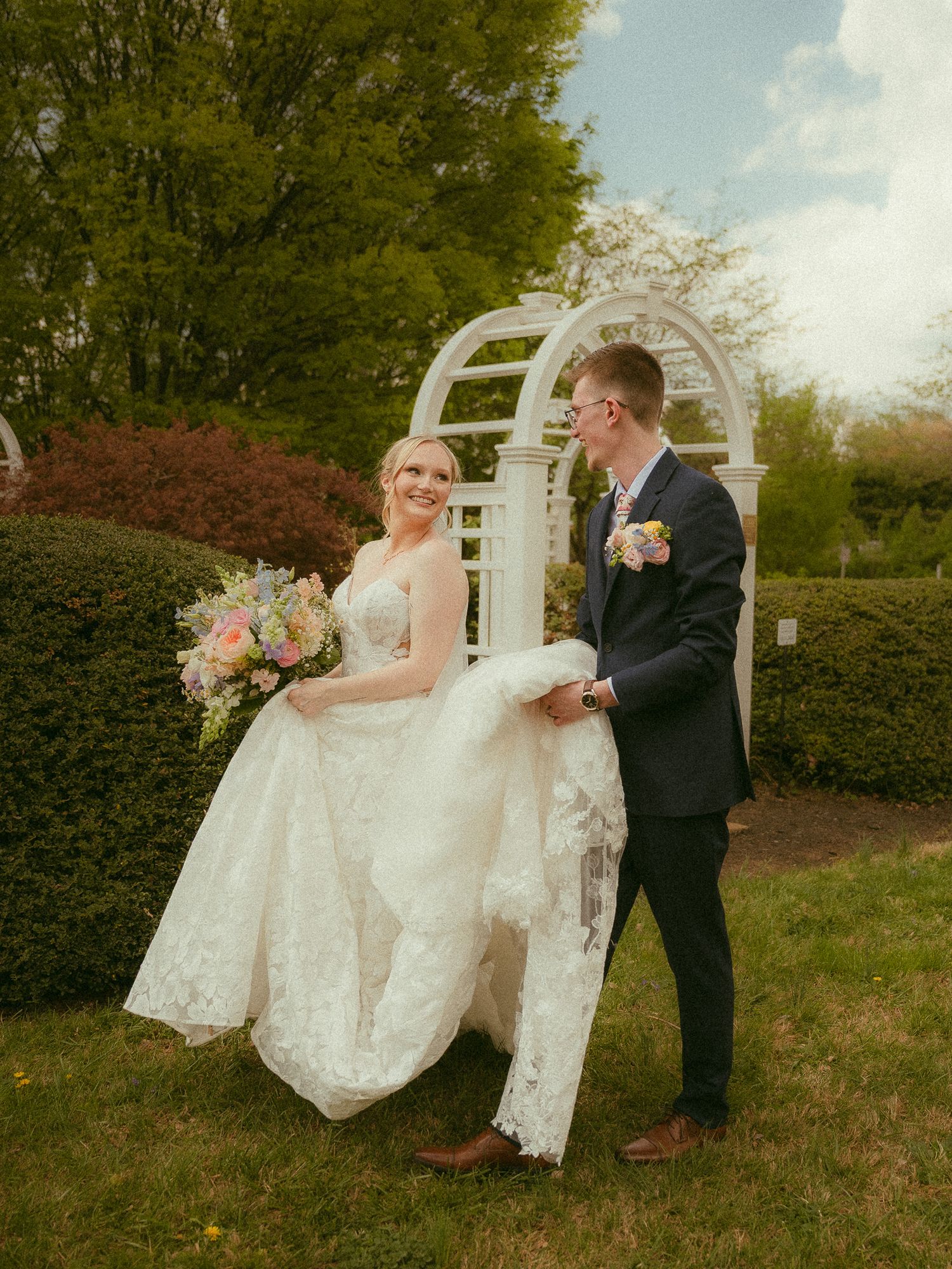 Bride and groom walk together in lush garden, bride holding colorful bouquet in white lace gown, groom in navy suit.