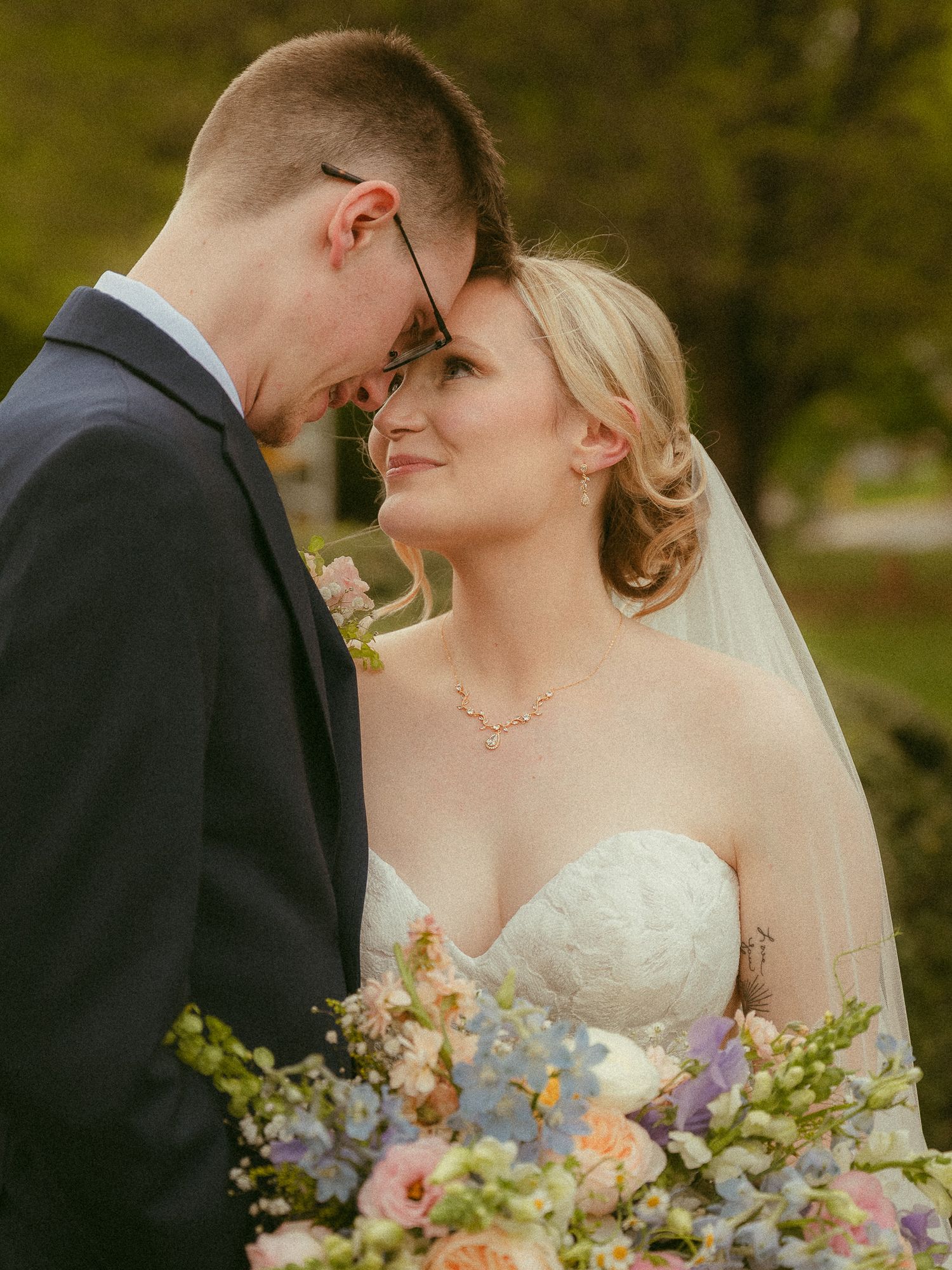 A bride and groom share an intimate forehead touch moment, bride holding pastel floral bouquet in soft outdoor light.
