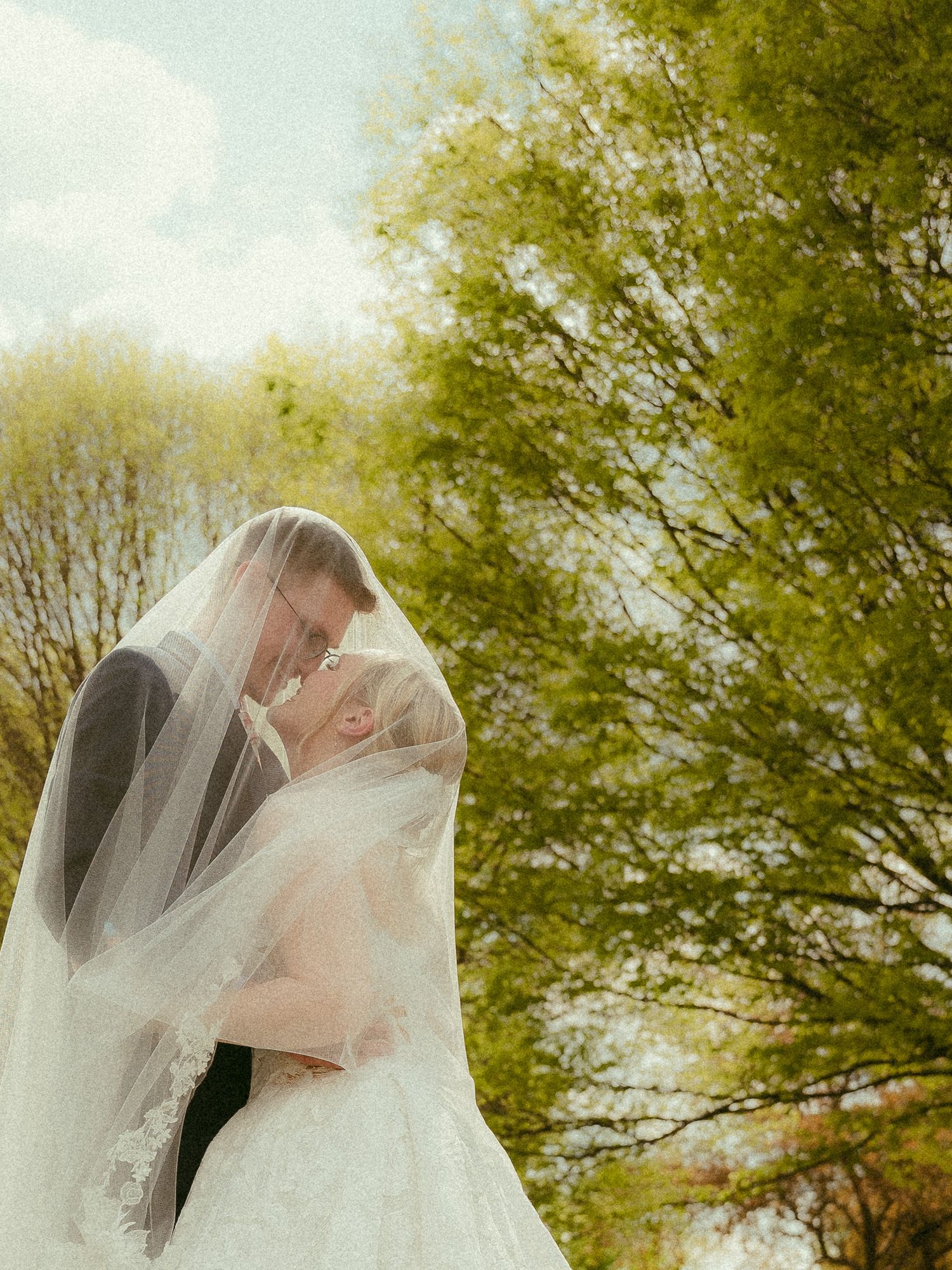 Bride and groom sharing a romantic kiss under a flowing white veil outdoors surrounded by lush green trees.