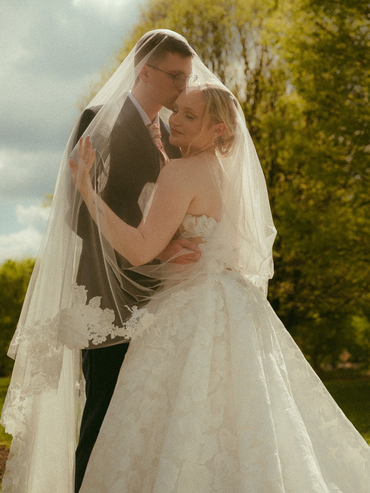 Bride and groom share romantic embrace outdoors, bride wearing lace wedding gown and veil, surrounded by lush green trees.