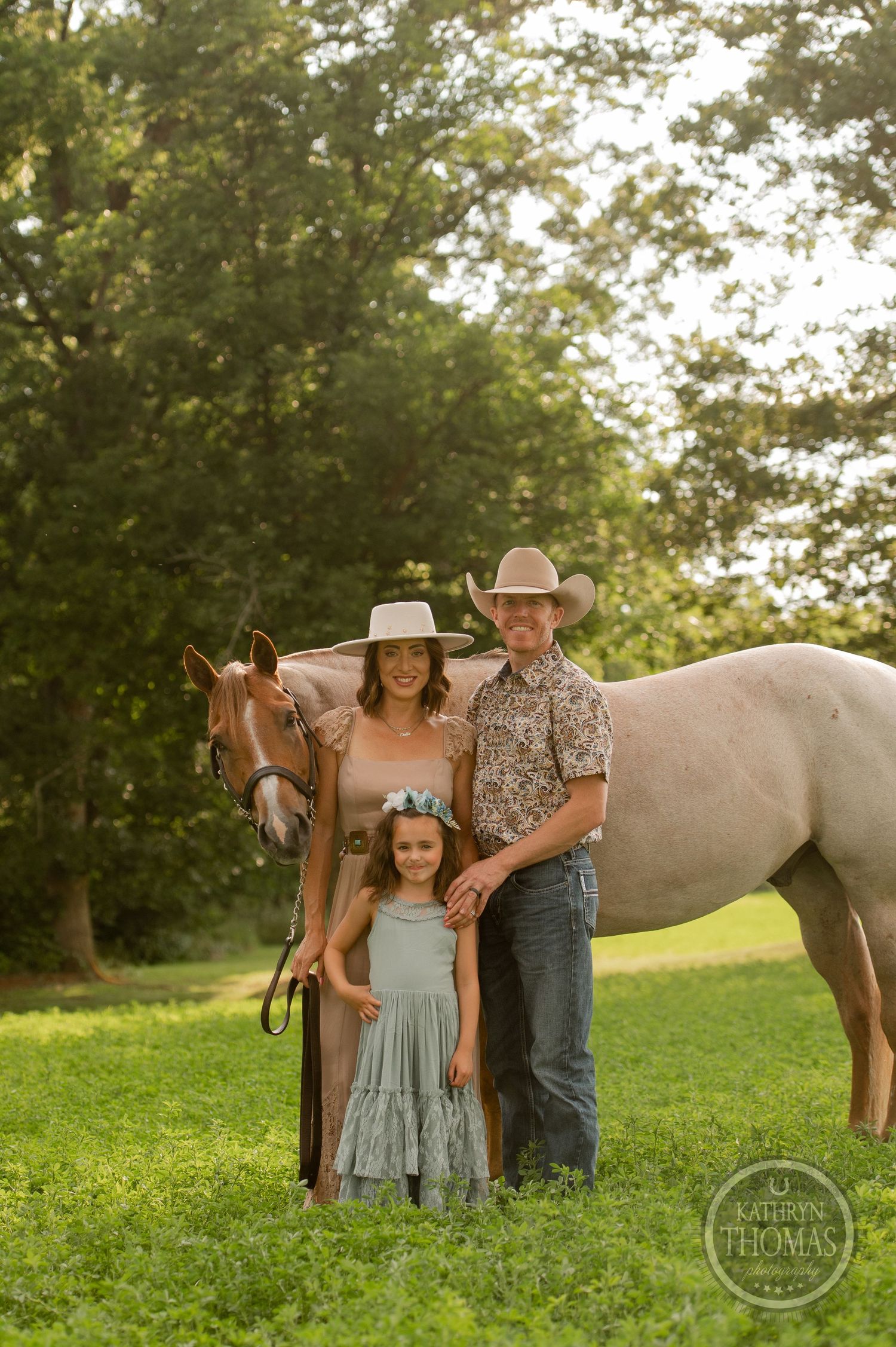 A family stands together with their pale horse in a sunlit meadow near a wooded area.