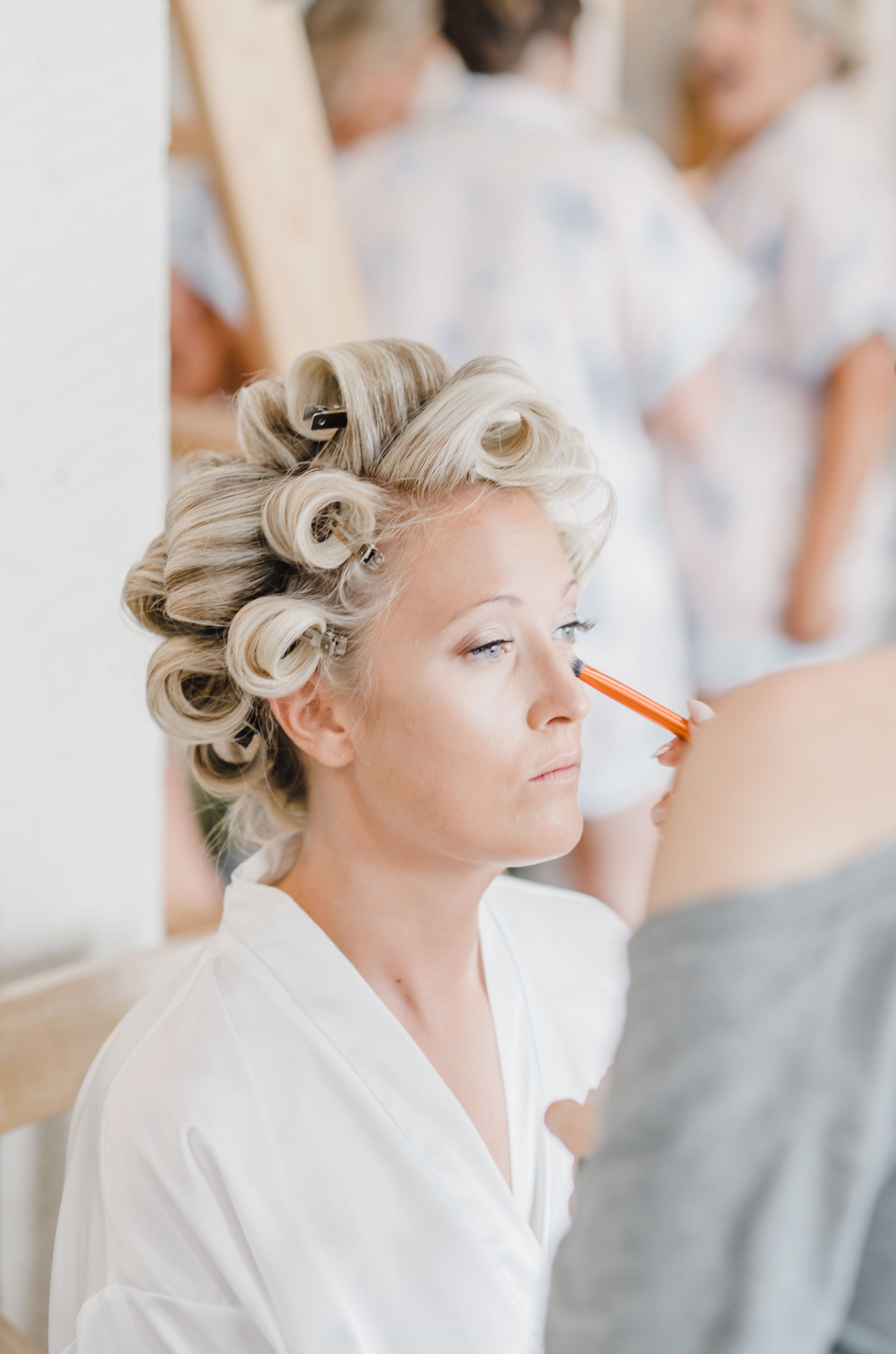 Wedding preparation photo showing vintage-inspired hairstyling with pin curls during makeup application.
