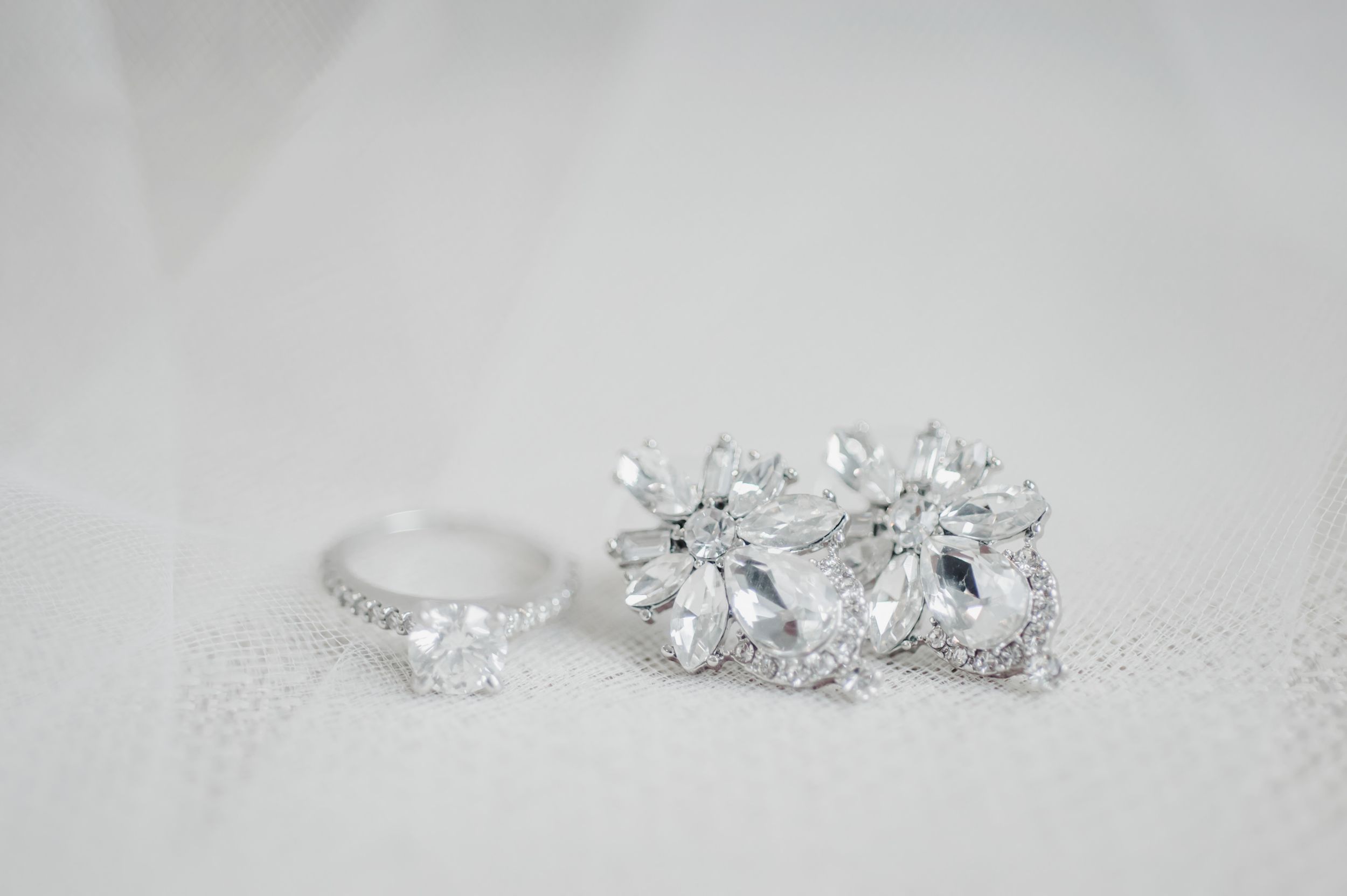 Sparkling diamond engagement ring and earrings arranged on white surface for wedding detail photography.