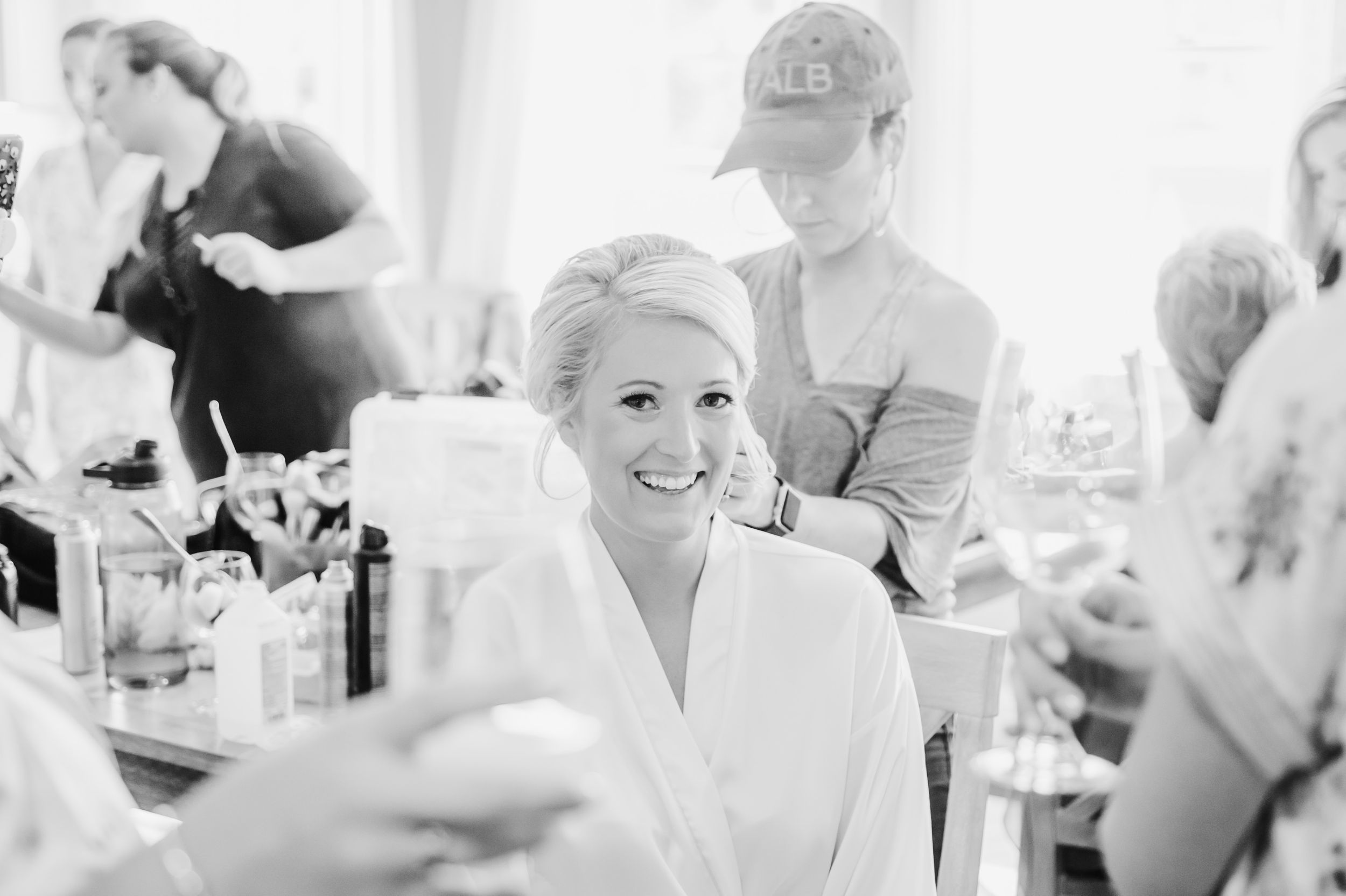 Black and white candid shot of pre-wedding preparations in a brightly lit room with makeup and styling.