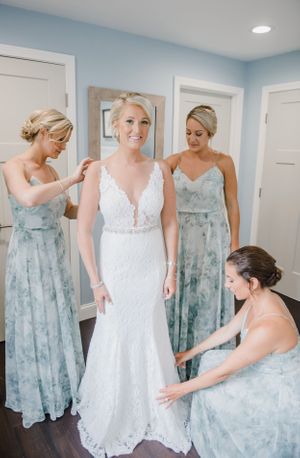 A bride in a white lace dress stands surrounded by bridesmaids wearing soft blue floral gowns during wedding preparations.
