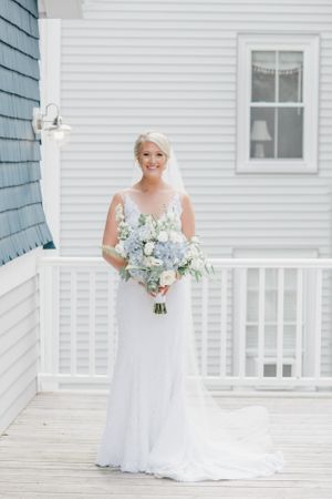 A bride in a white gown poses with her bouquet on a wooden porch of a coastal home.