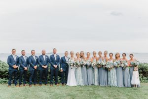 Wedding party stands in formal arrangement with ocean view and cloudy sky backdrop.