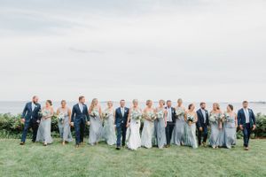 Full wedding party in navy and light blue attire poses on oceanfront lawn.