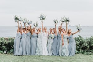 Bridesmaids in light blue dresses raise bouquets in celebration on coastal lawn.